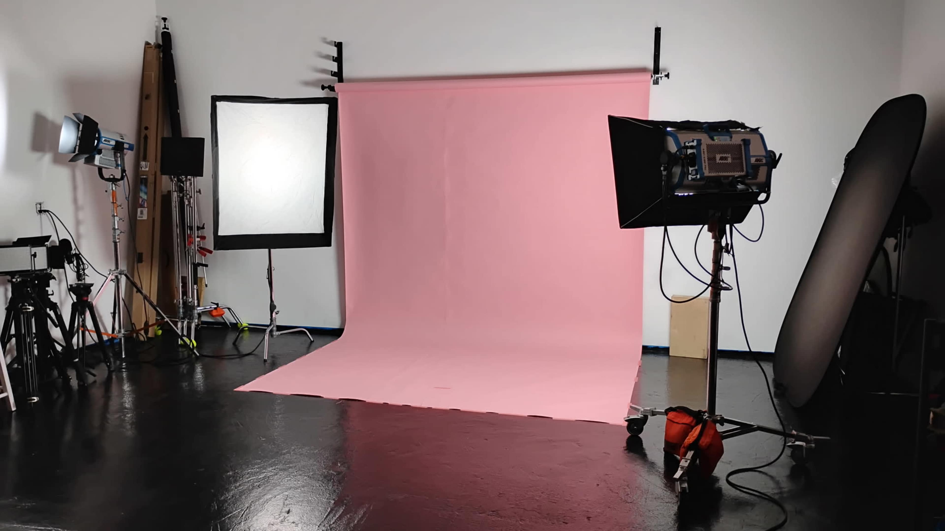 The image shows a photography studio featuring a pink backdrop, studio lights, and various equipment arranged around the scene.