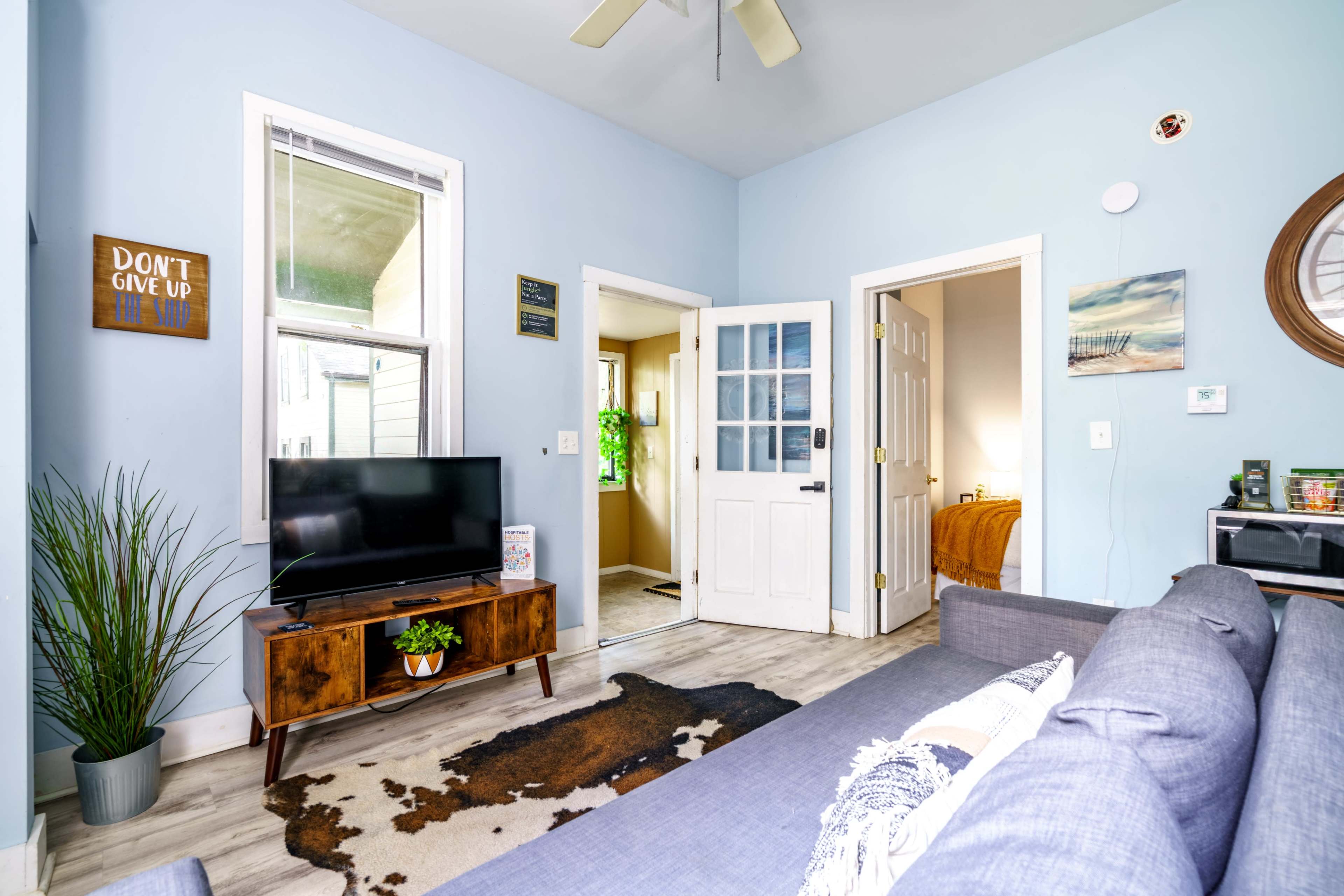 The image shows a cozy living room with a blue wall, a gray couch, a TV on a wooden stand, and a door leading to another room.