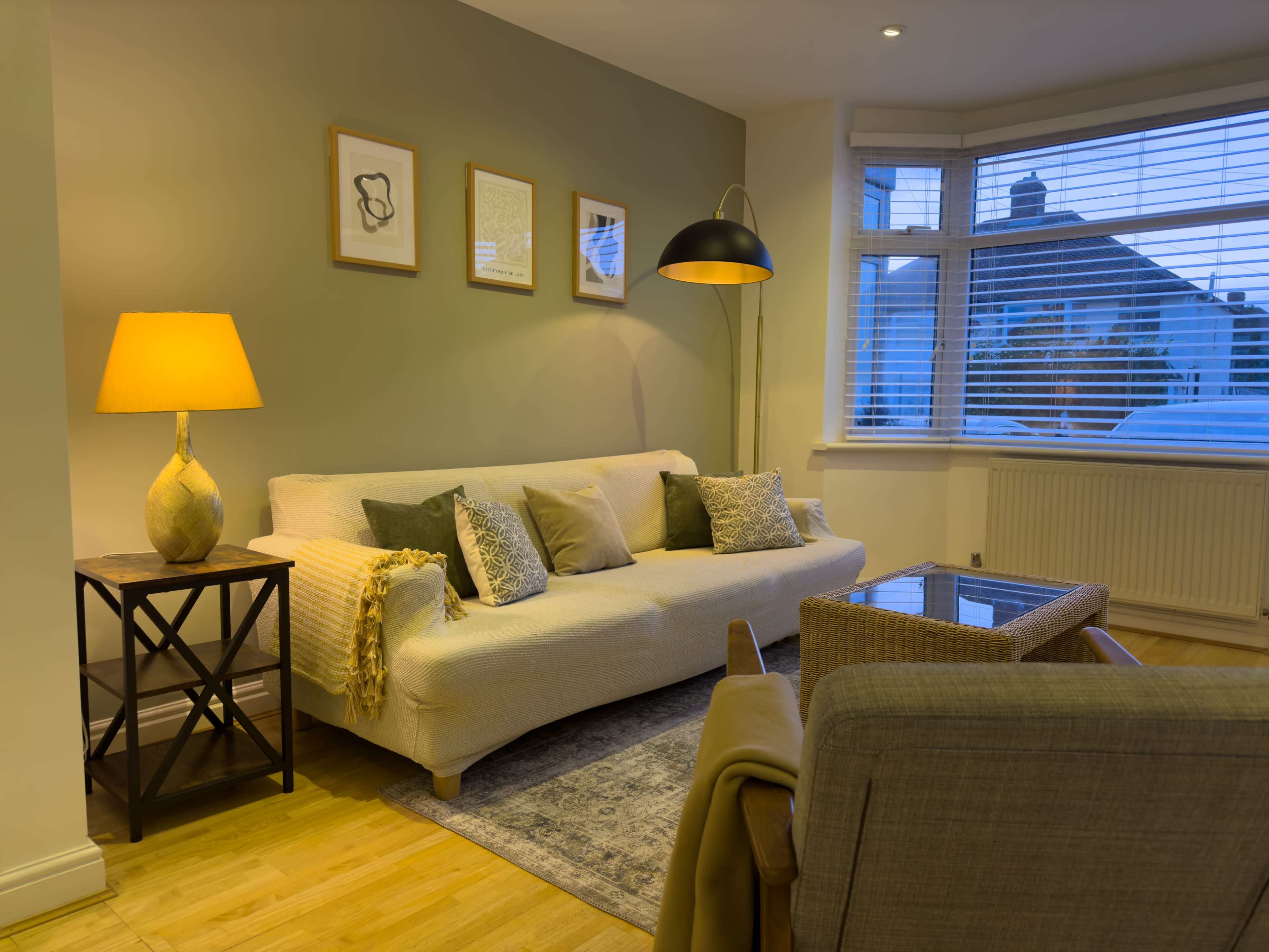 A cozy living room with a light-colored sofa, a wooden side table with a lamp, and a large window with white blinds.