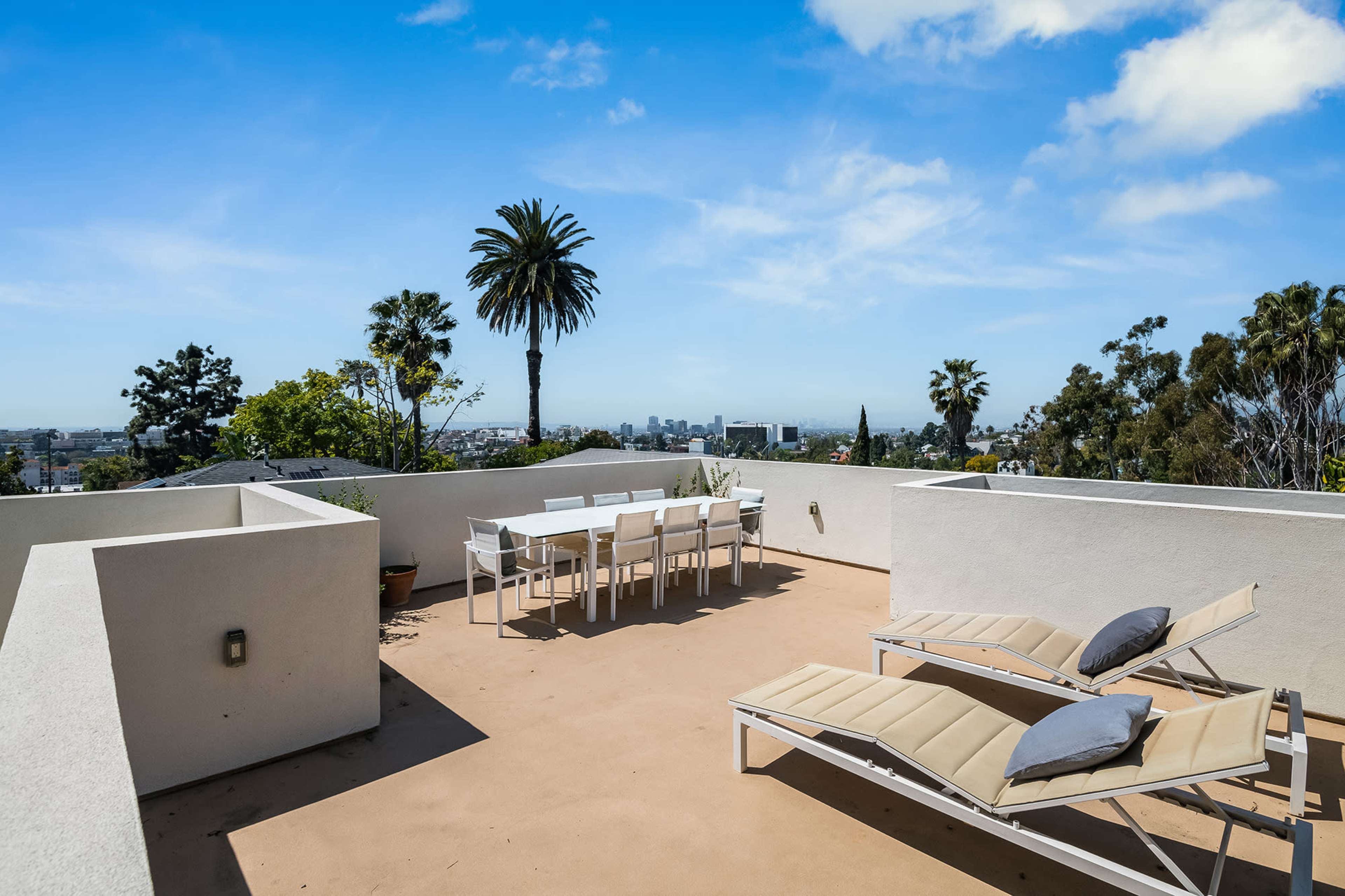 The image shows a rooftop terrace with white patio furniture, including a dining table and lounge chairs, overlooking a cityscape with palm trees in the background.