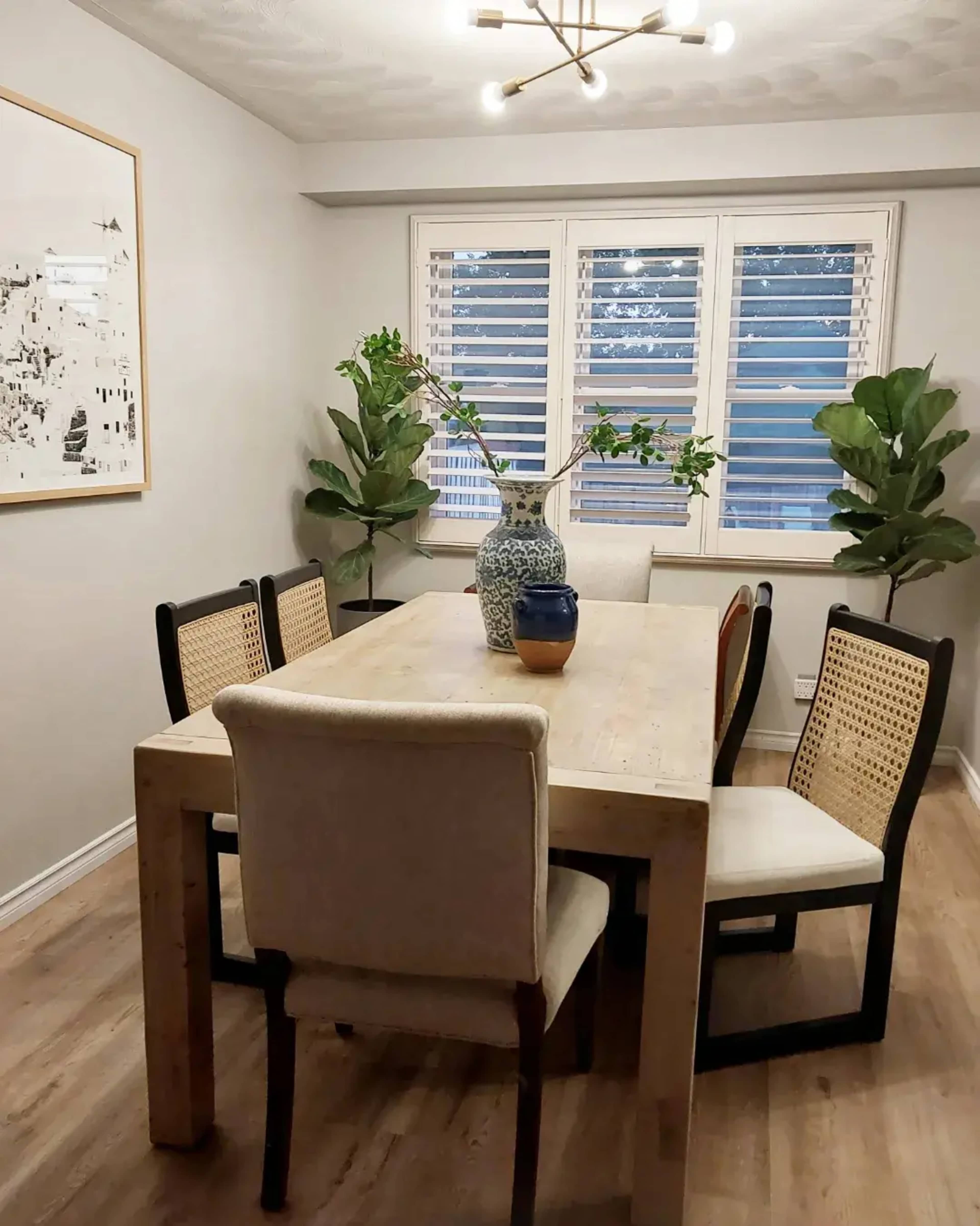 A dining room features a wooden table surrounded by six chairs, with large plants and a vase on the table, beneath a modern light fixture.