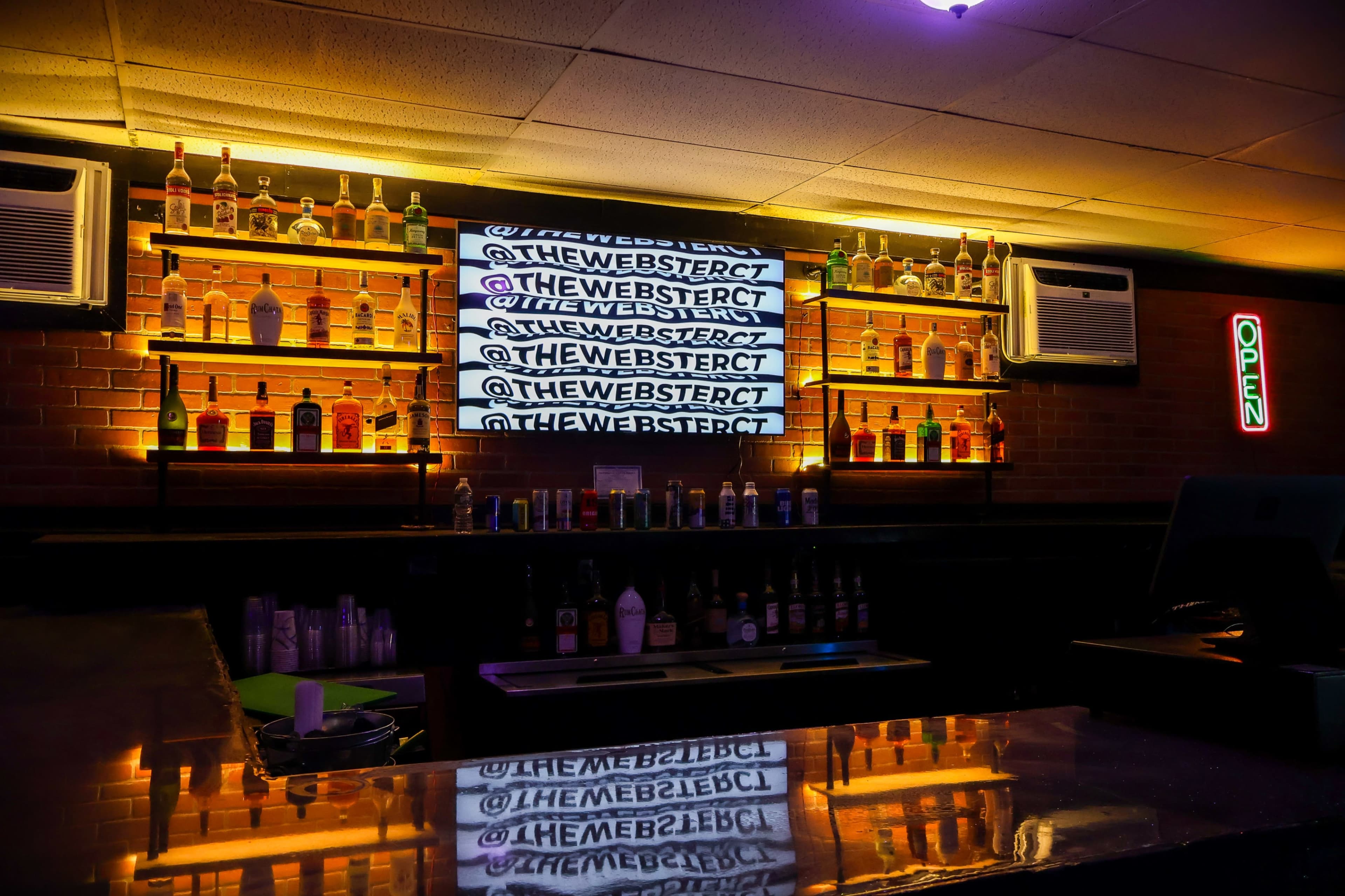 The image shows a bar interior with shelves displaying various bottles, a large screen advertising the bar's social media handles, and an "OPEN" sign illuminated on the wall.