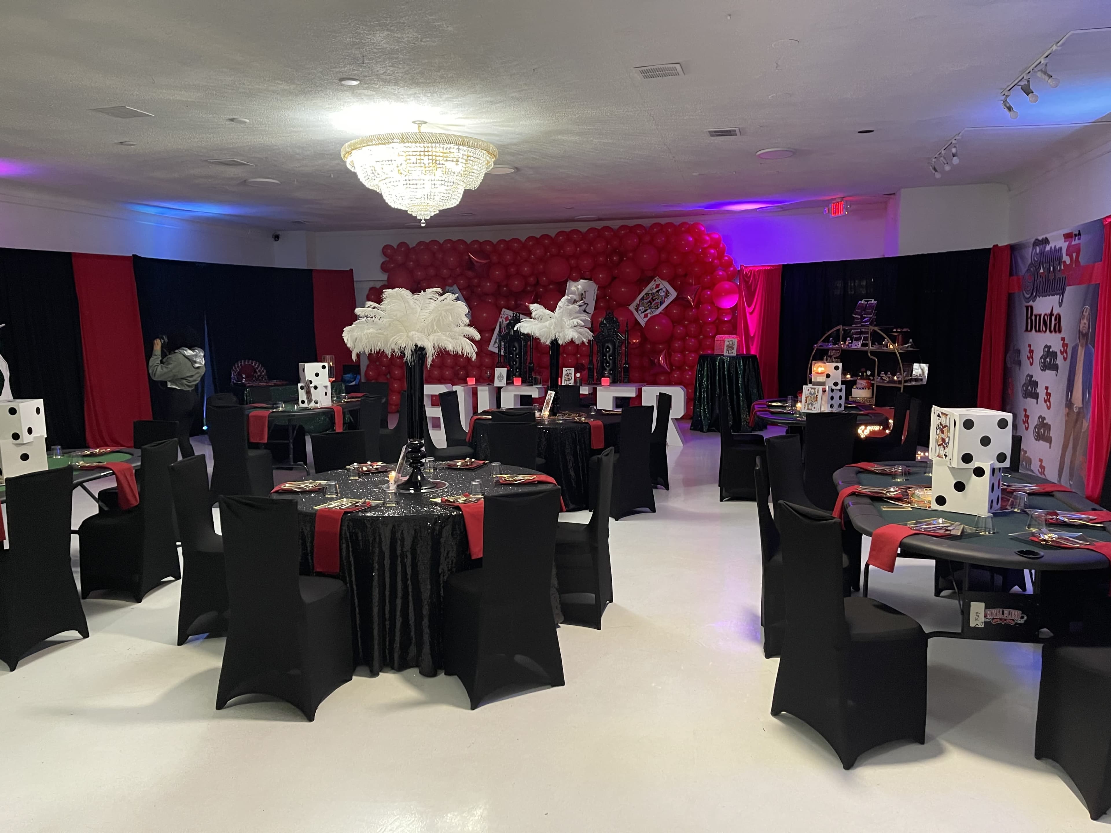 The room is set up for an event, featuring black and red tables adorned with decorations, large dice, and feather centerpieces, surrounded by balloon arches.