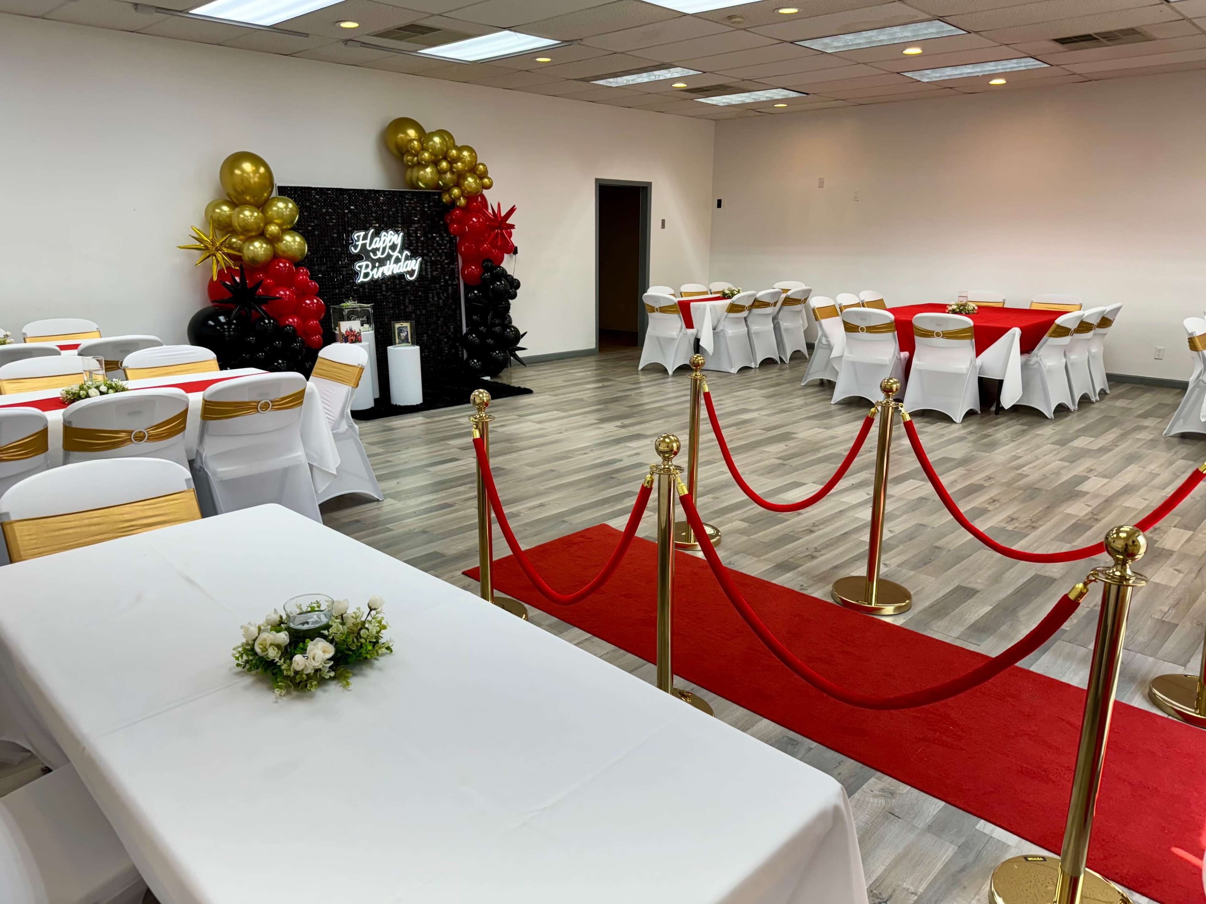A decorated event space featuring a red carpet entrance, gold and black balloons, and tables set for a birthday celebration.