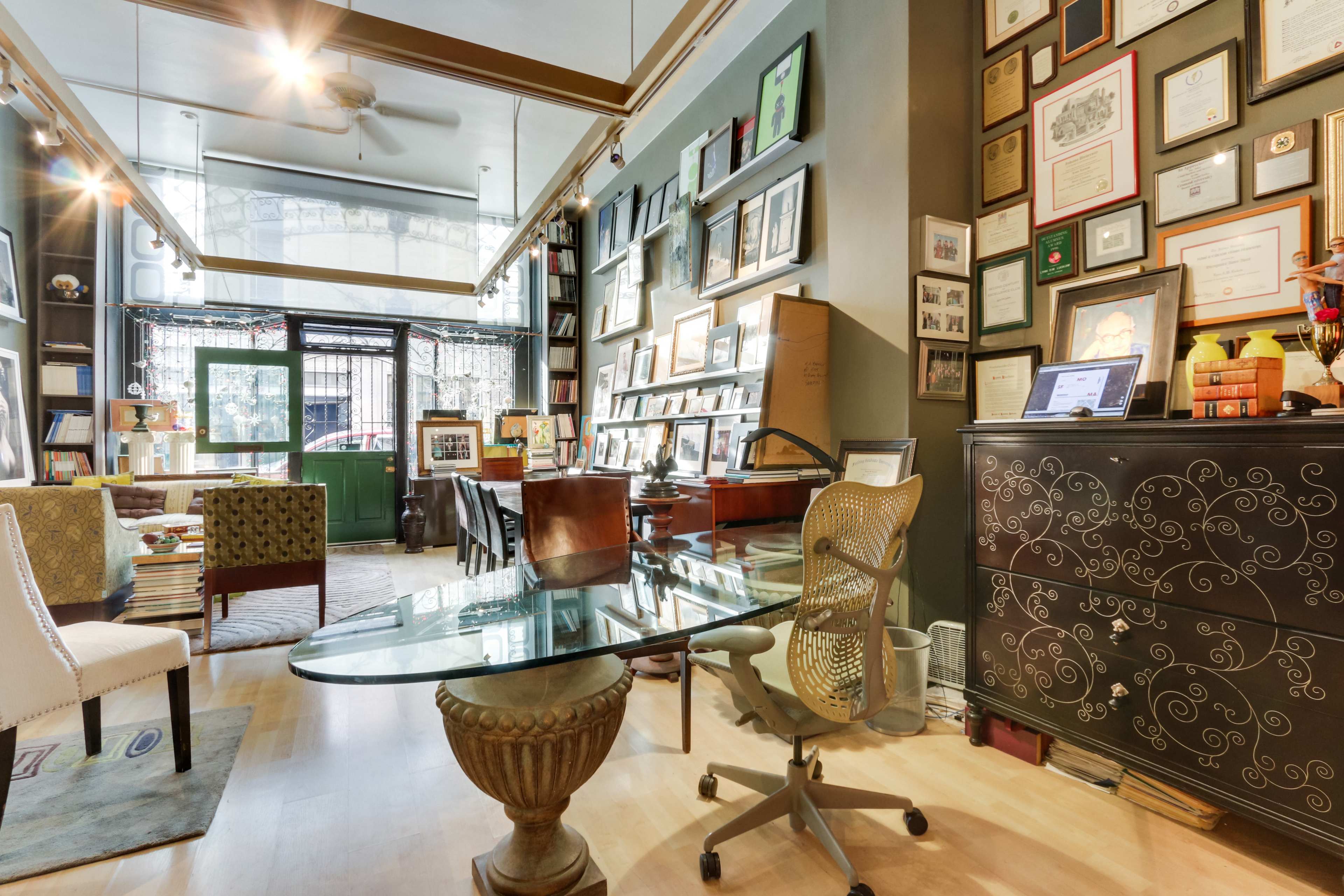 A cozy office space with a glass desk, wooden chairs, and a wall adorned with various framed certificates and photographs.