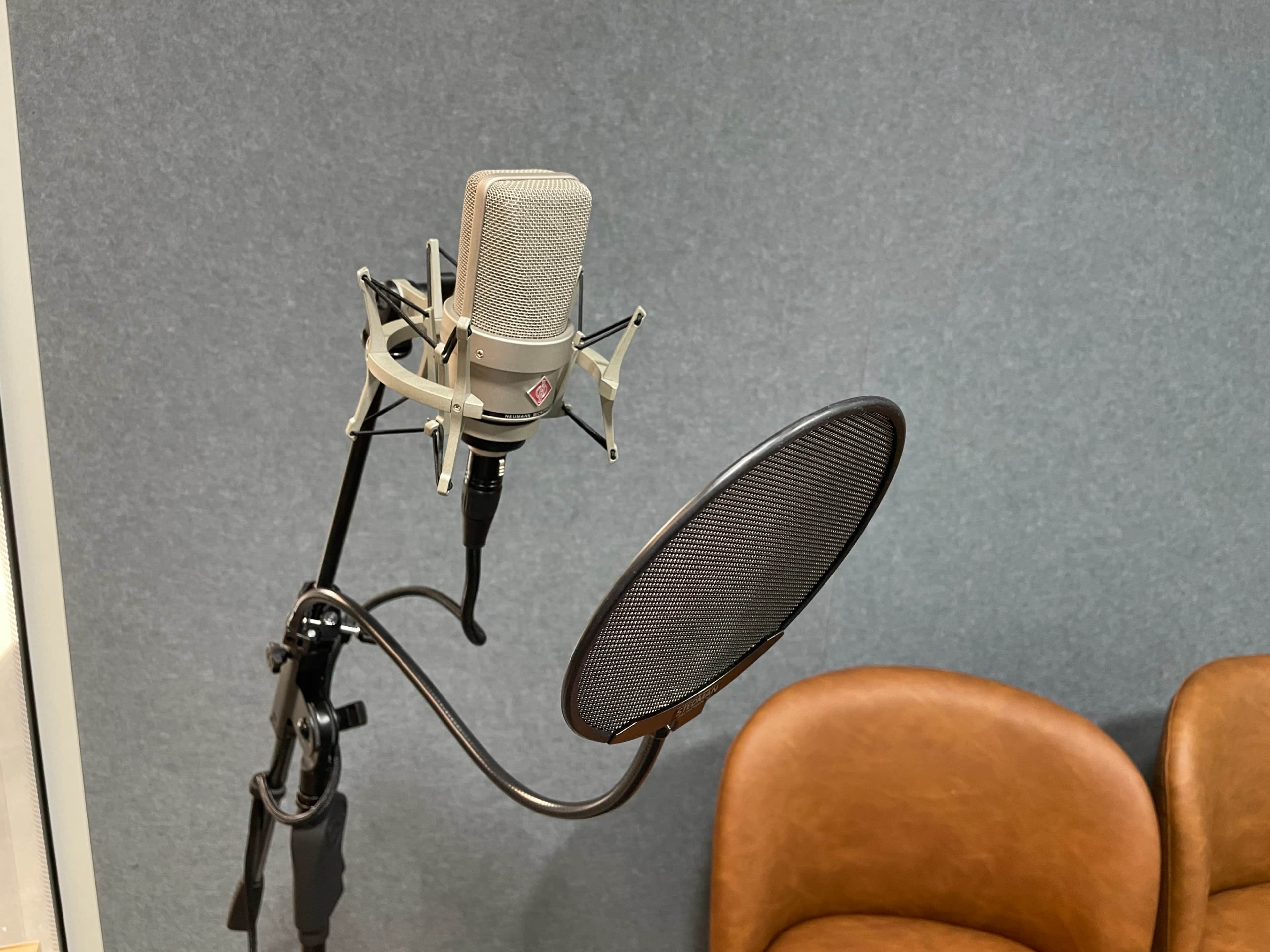 A microphone on a stand is positioned next to a pop filter, with two brown chairs in the background against a gray wall.