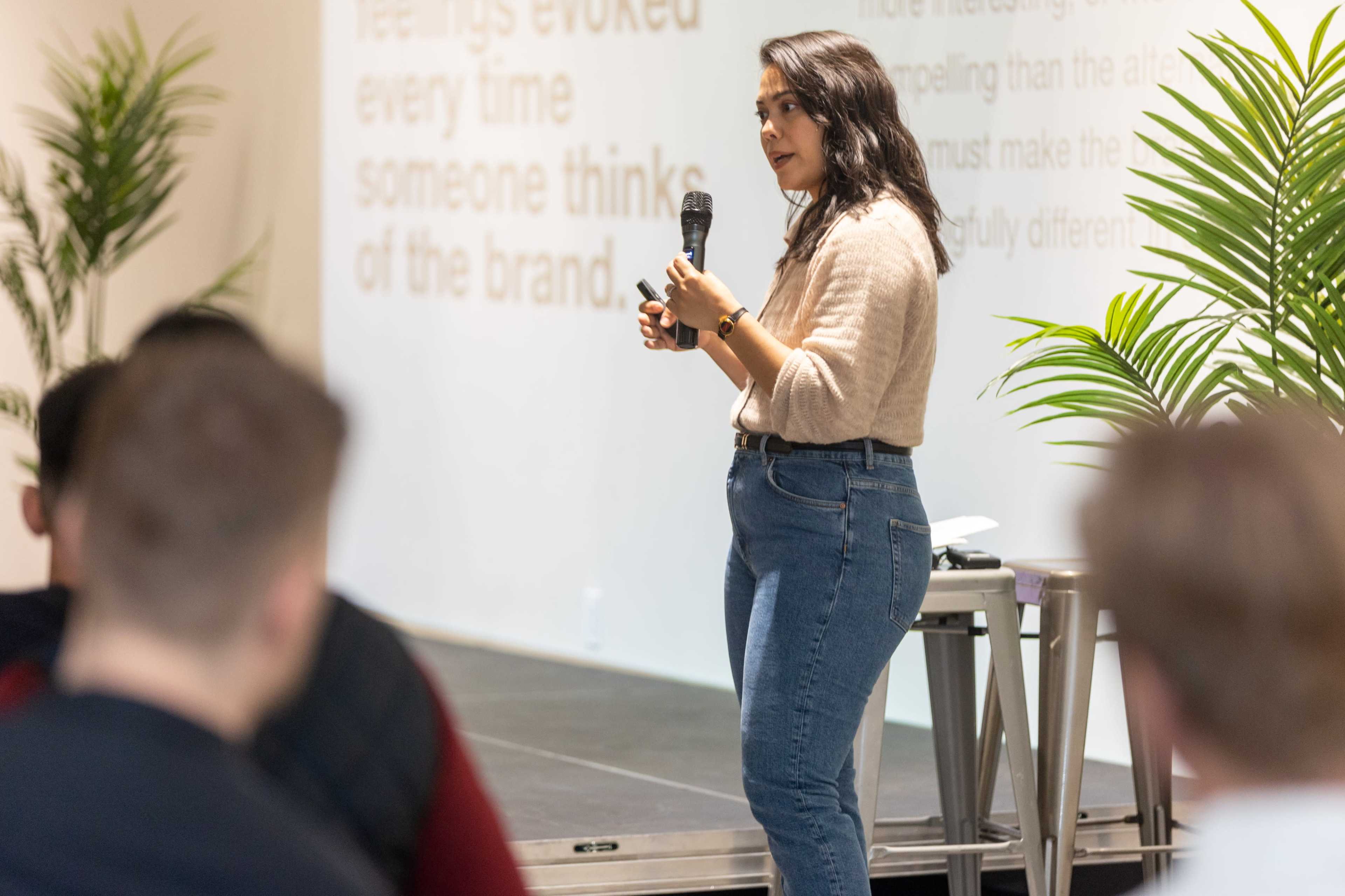 A woman stands on stage holding a microphone while speaking to an audience seated in front of her.