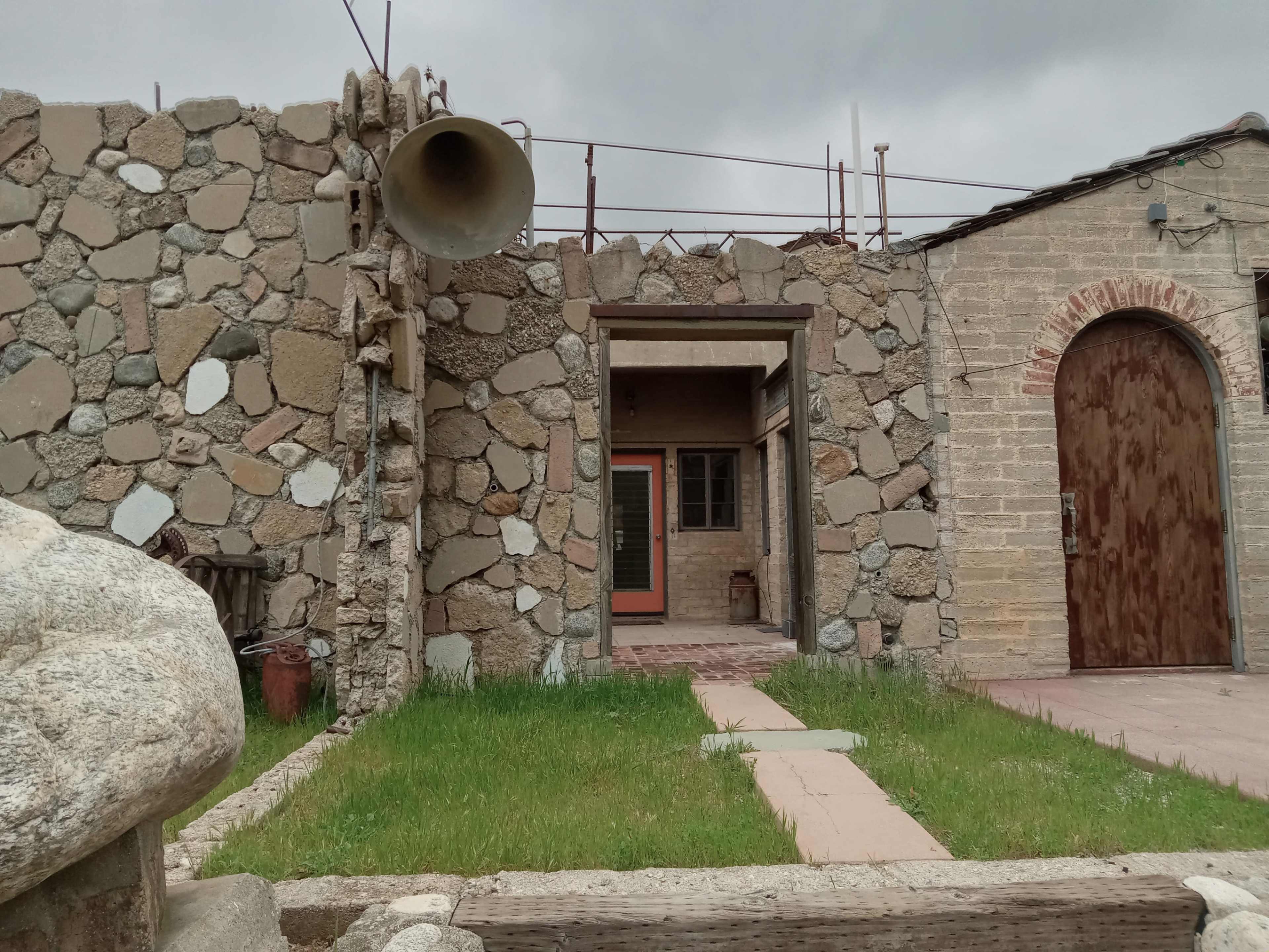 The image shows a stone-walled structure with a large horn speaker mounted on the side, leading to an entrance door and visible pathways in a grassy area.