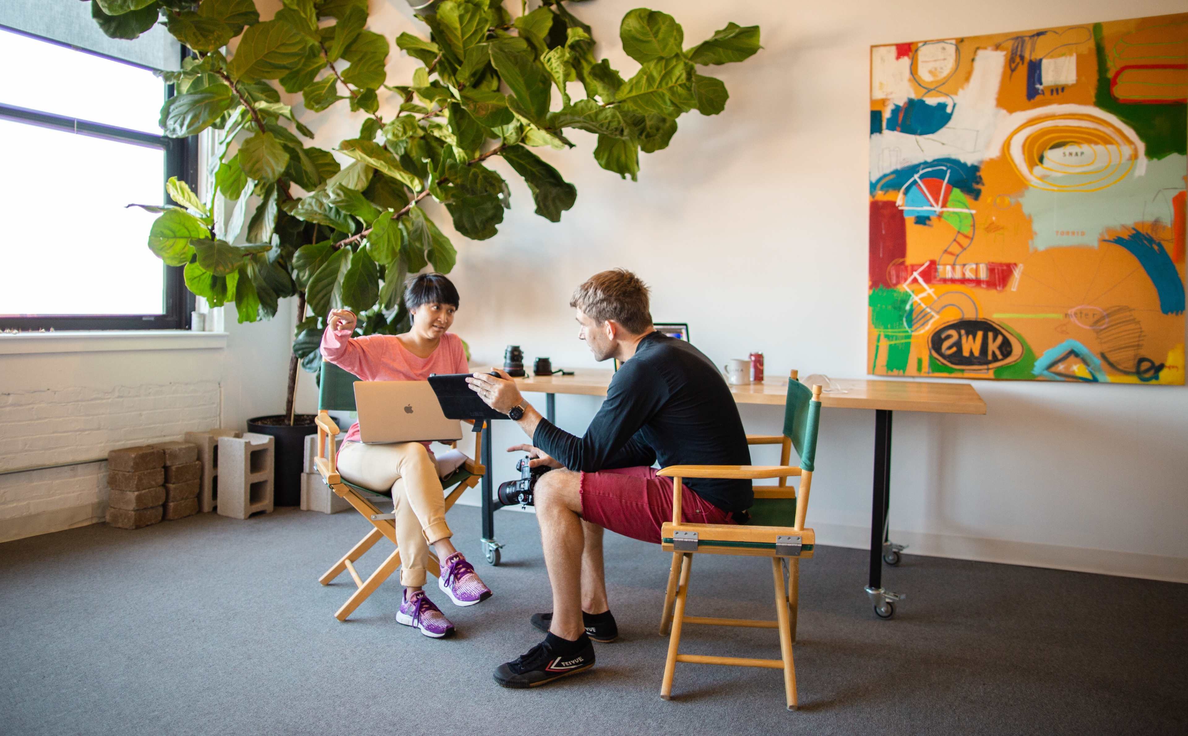 A woman and a man are seated in a brightly lit room, discussing while facing each other with a laptop open on a small table between them.