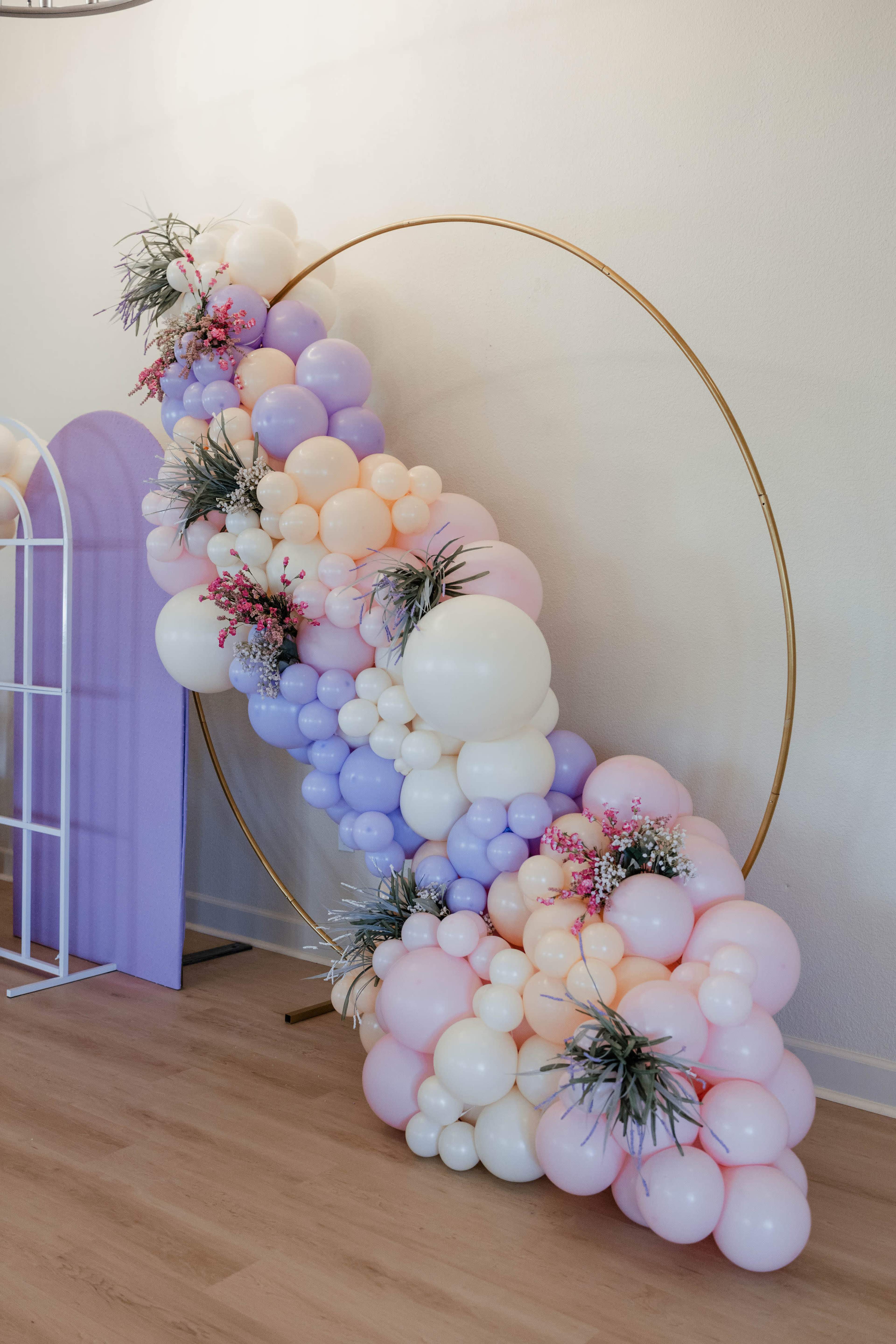 A large circular frame adorned with pastel-colored balloons and floral accents is displayed against a plain wall.