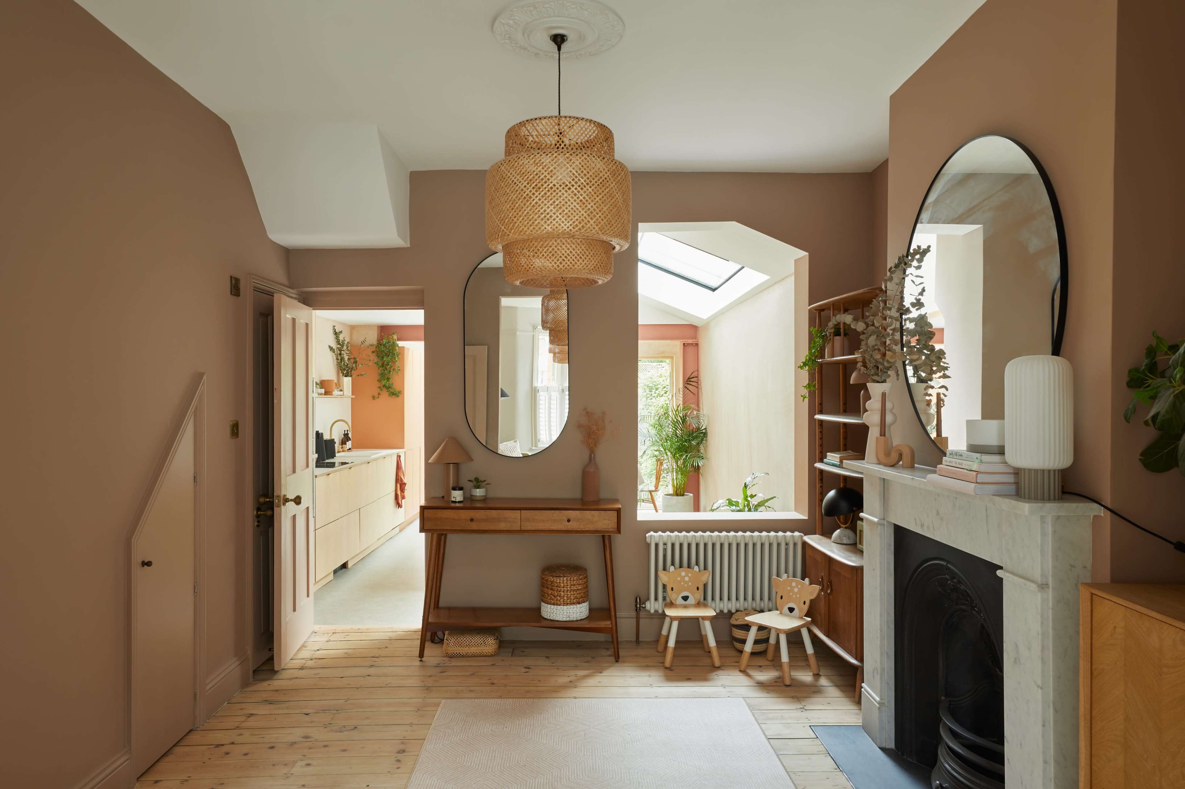 A warm-toned hallway with a wooden floor, featuring a round mirror above a console table, two small chairs, and a cozy seating area with plants.