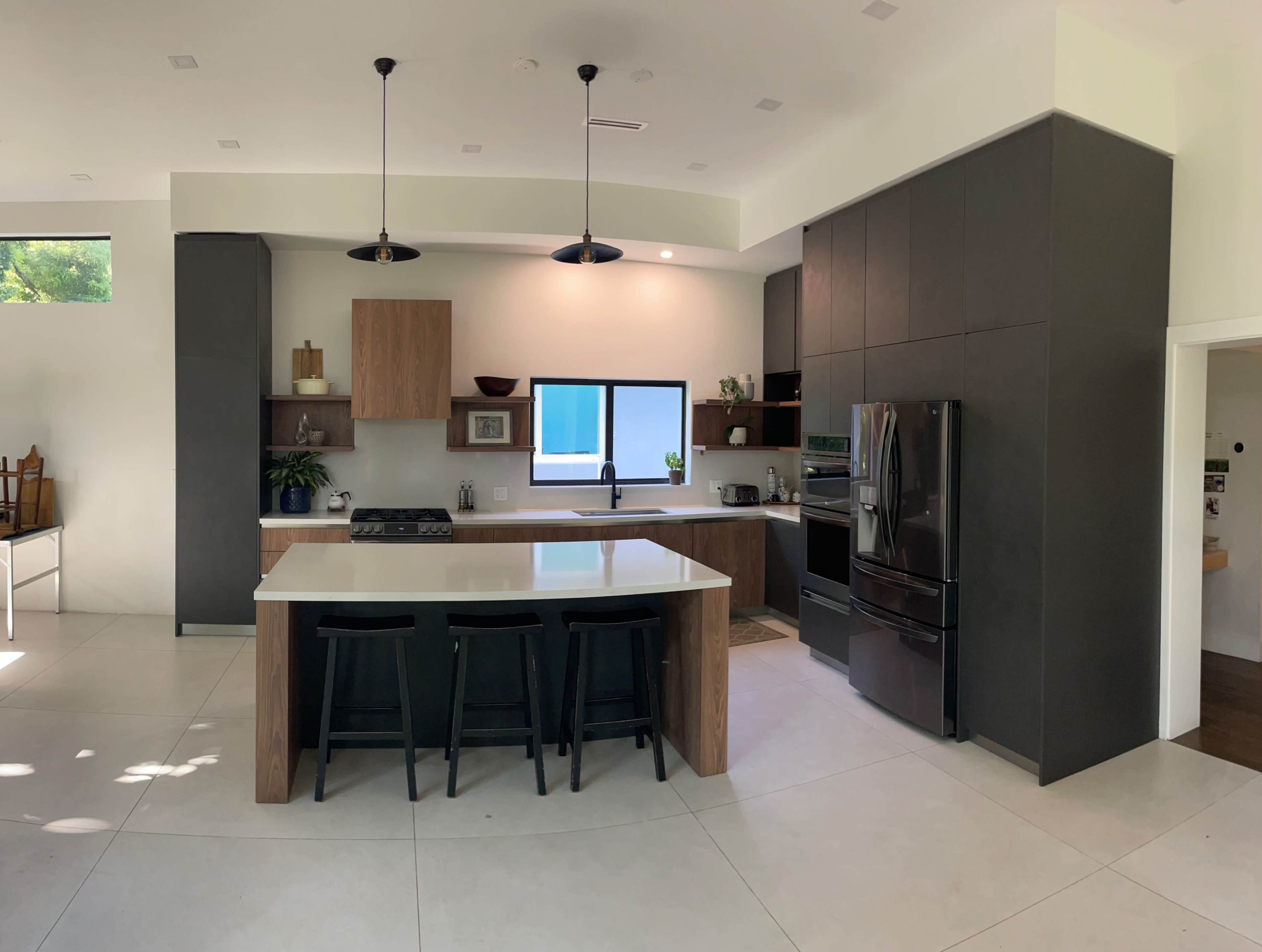 The image shows a modern kitchen featuring a central island with seating, dark cabinetry, stainless steel appliances, and a large window that allows natural light to enter the space.