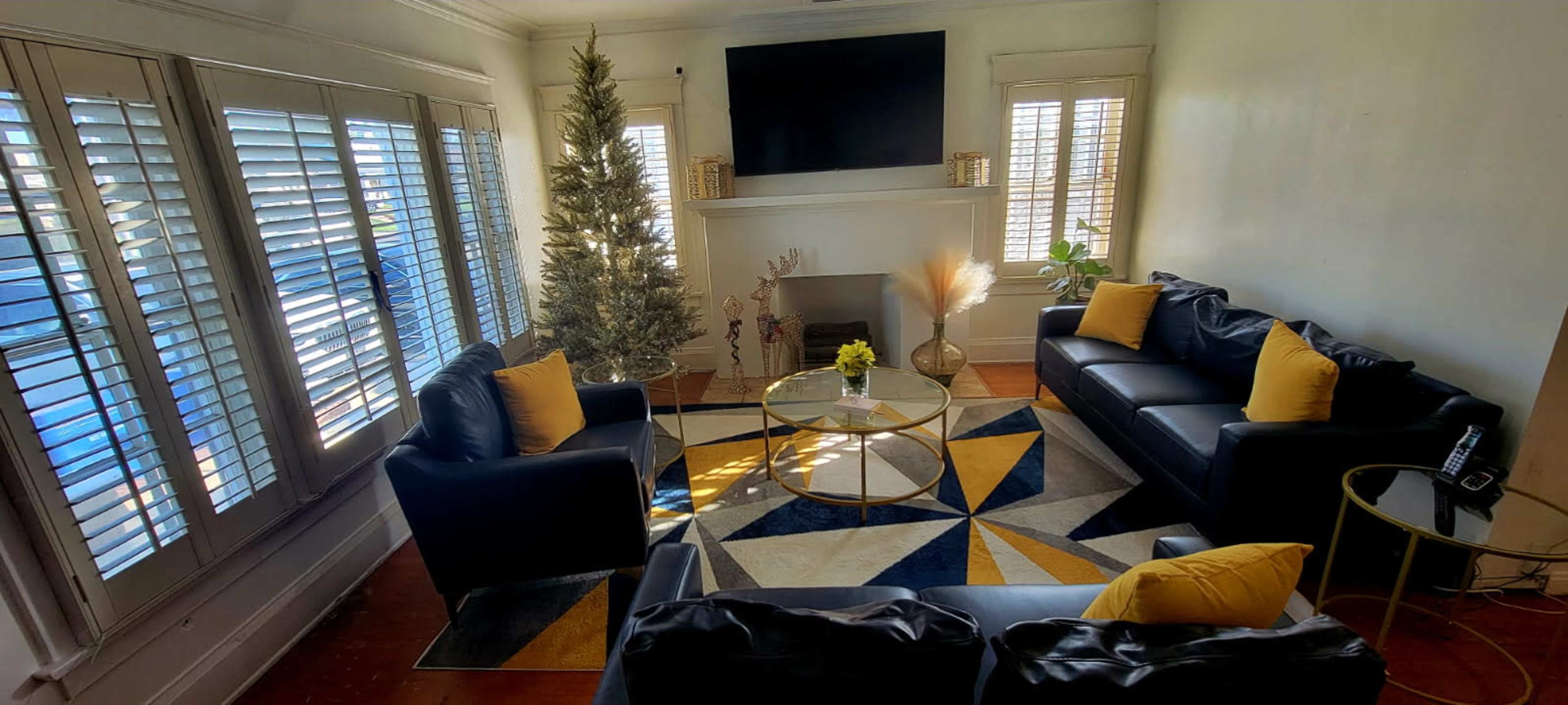 The image shows a living room with two black leather sofas, a glass coffee table, a yellow and blue geometric rug, and a decorative tree near a white fireplace.