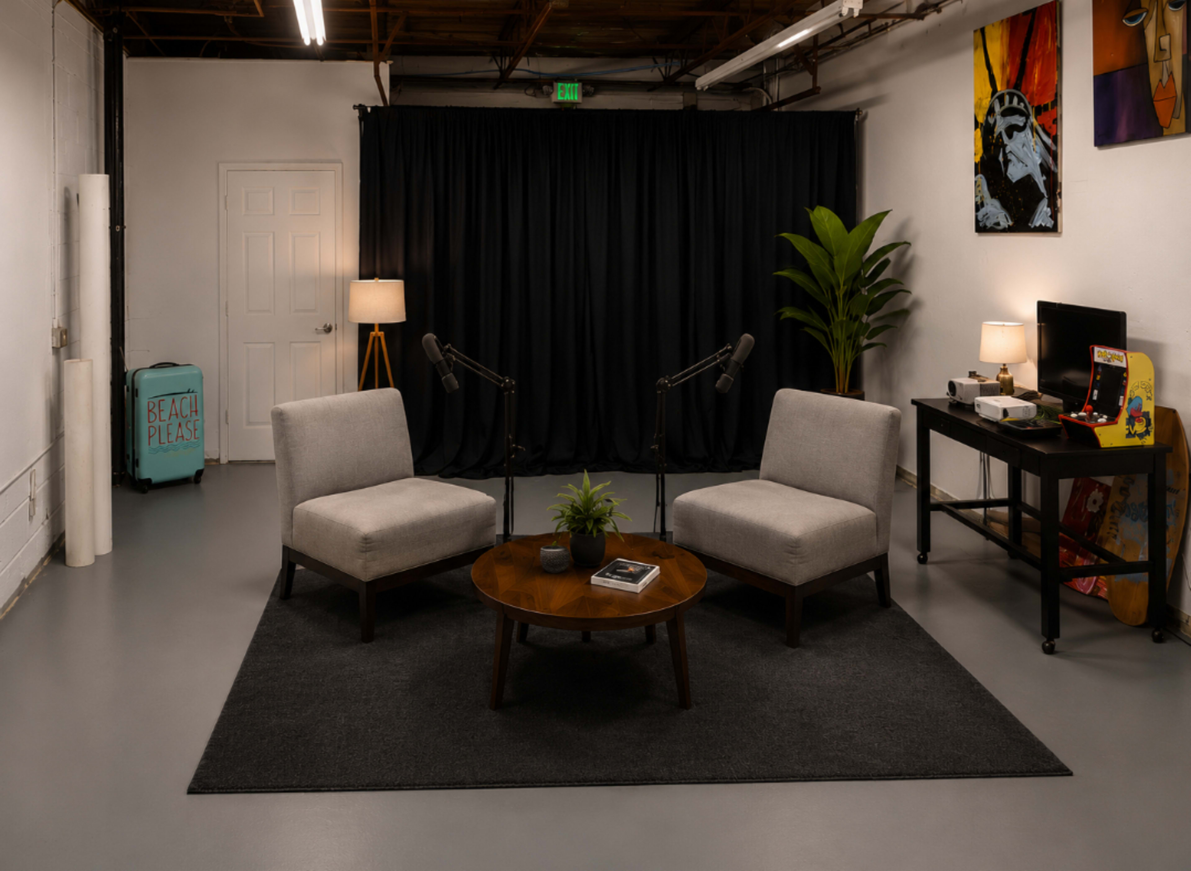 The image shows a minimalist room with two gray chairs facing a round table, surrounded by a black curtain, a plant, and various furnishings like a lamp and a desk.