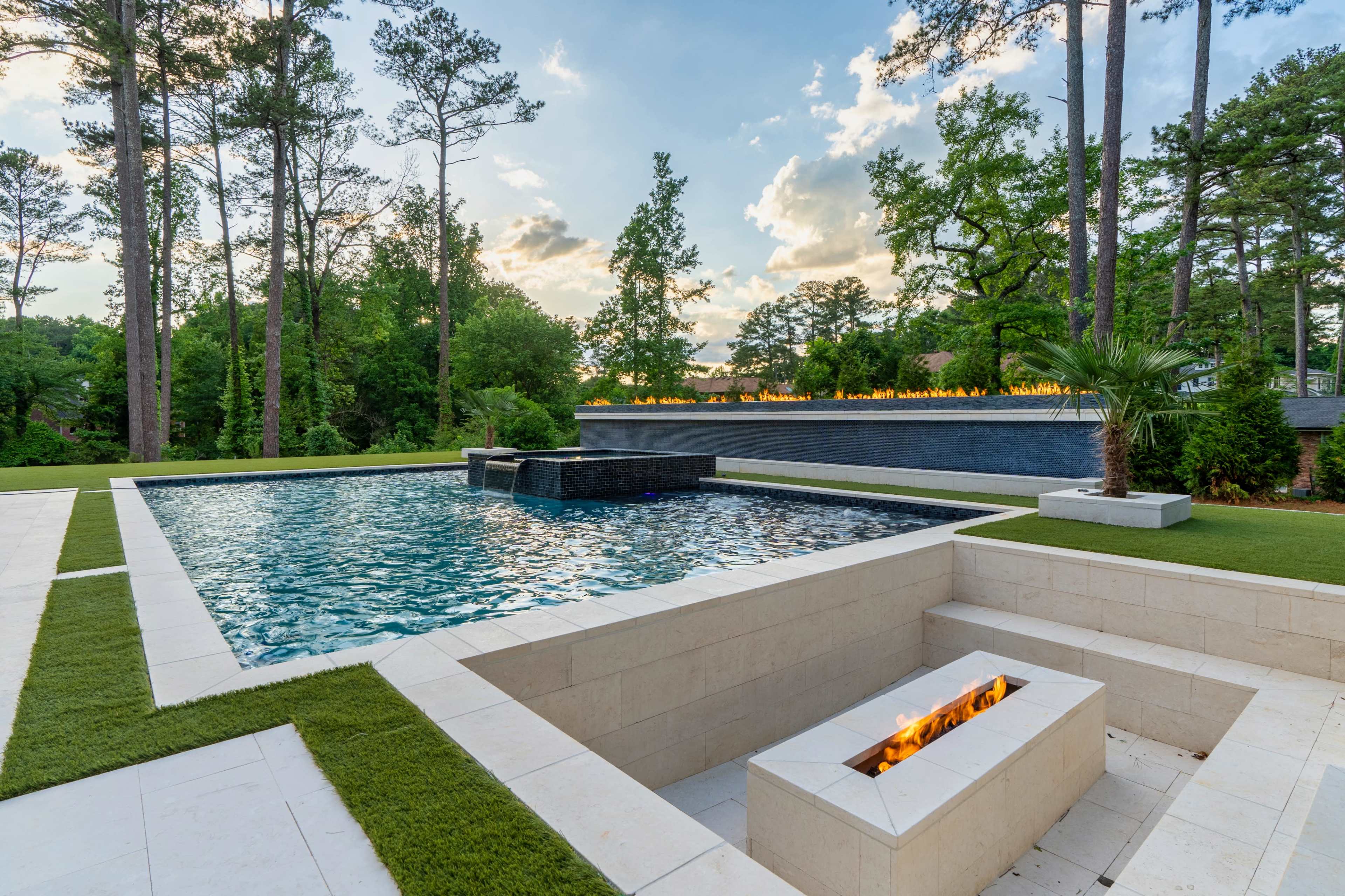 A modern outdoor pool area with a fire feature along the edge and lush greenery in the background.