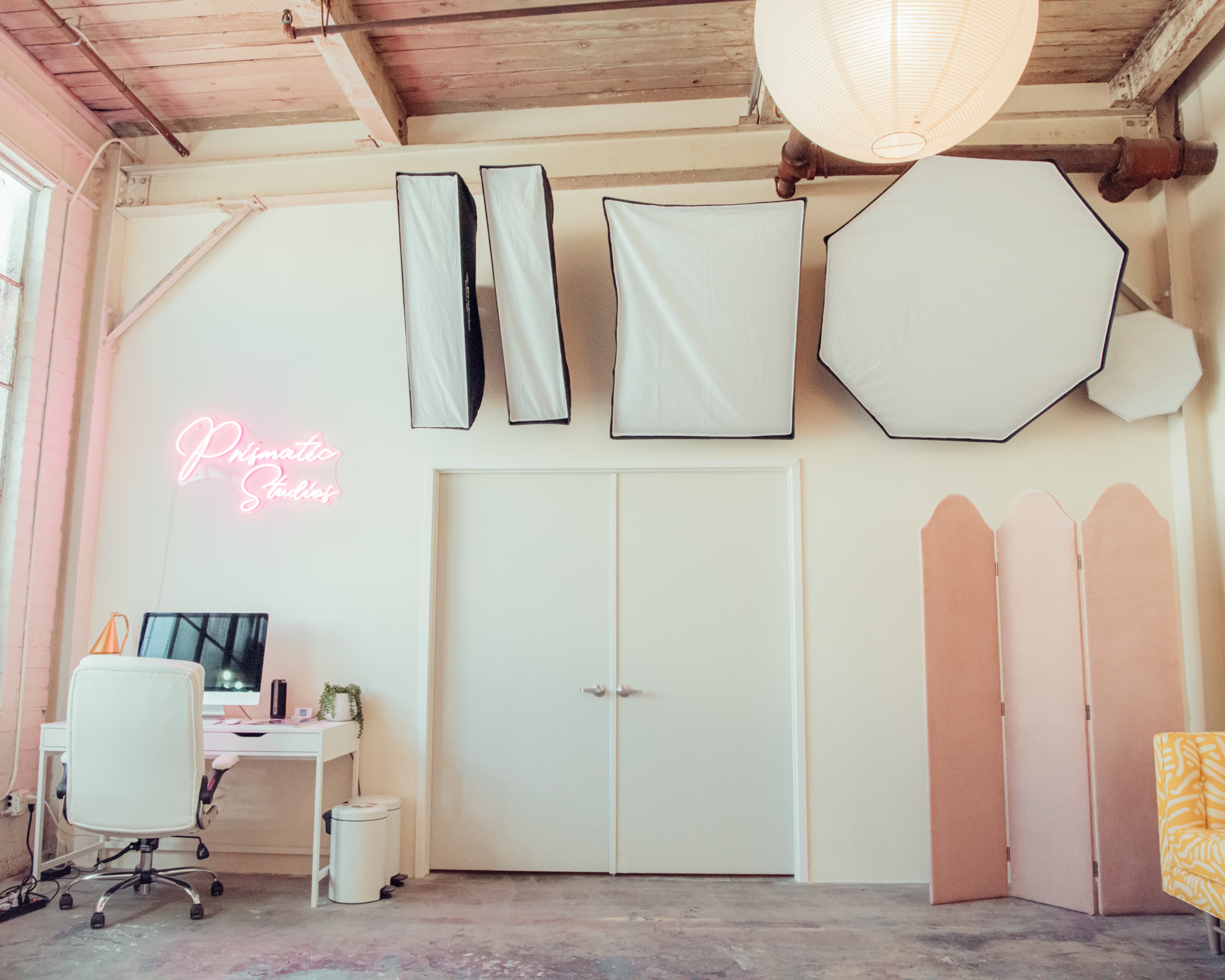The image shows a bright photography studio featuring a workspace with a desk and computer, along with large softboxes and a decorative pink partition on the wall.