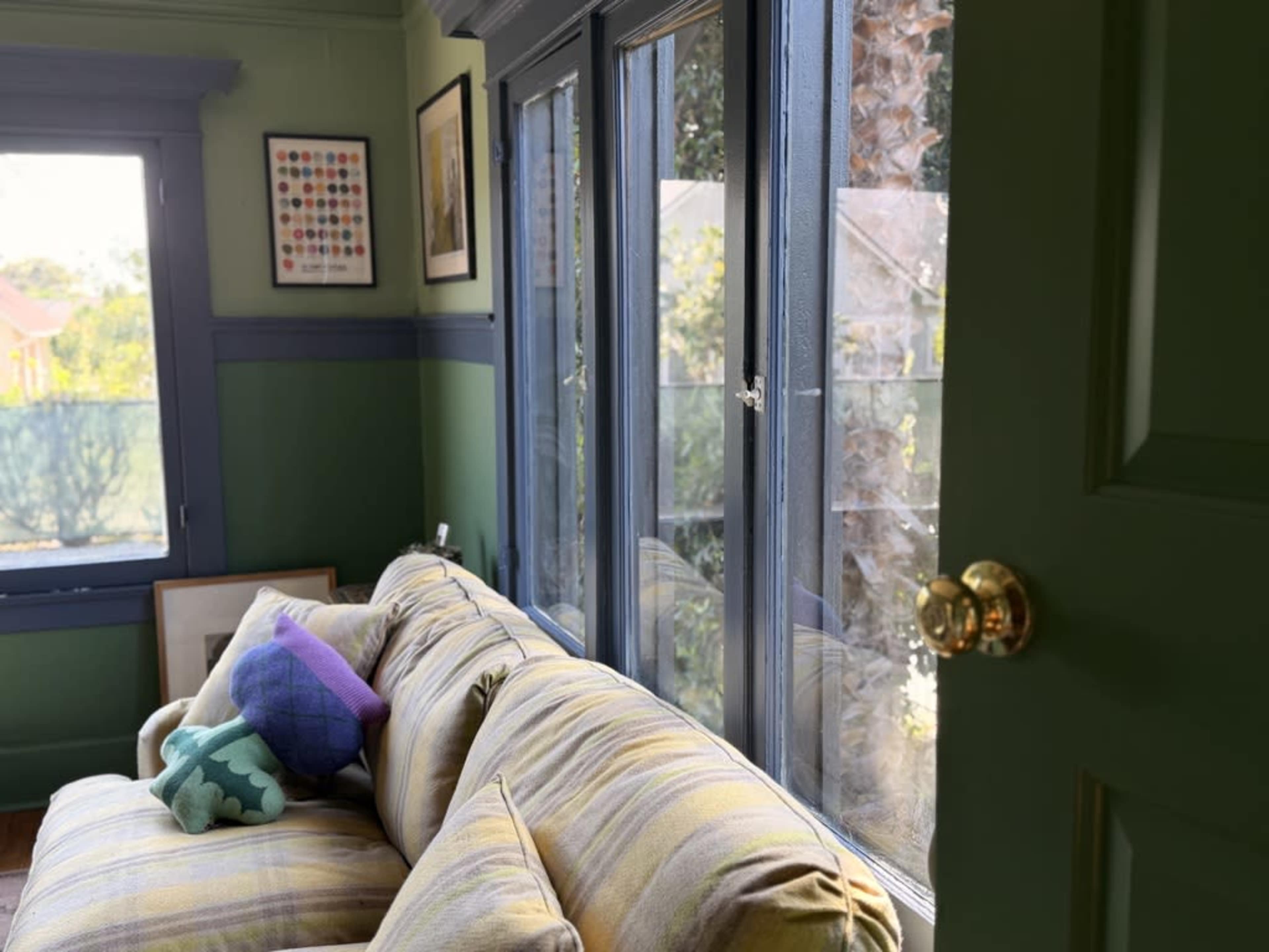 A sofa with a patterned throw pillow sits near large windows in a room with green walls and a door partially open.