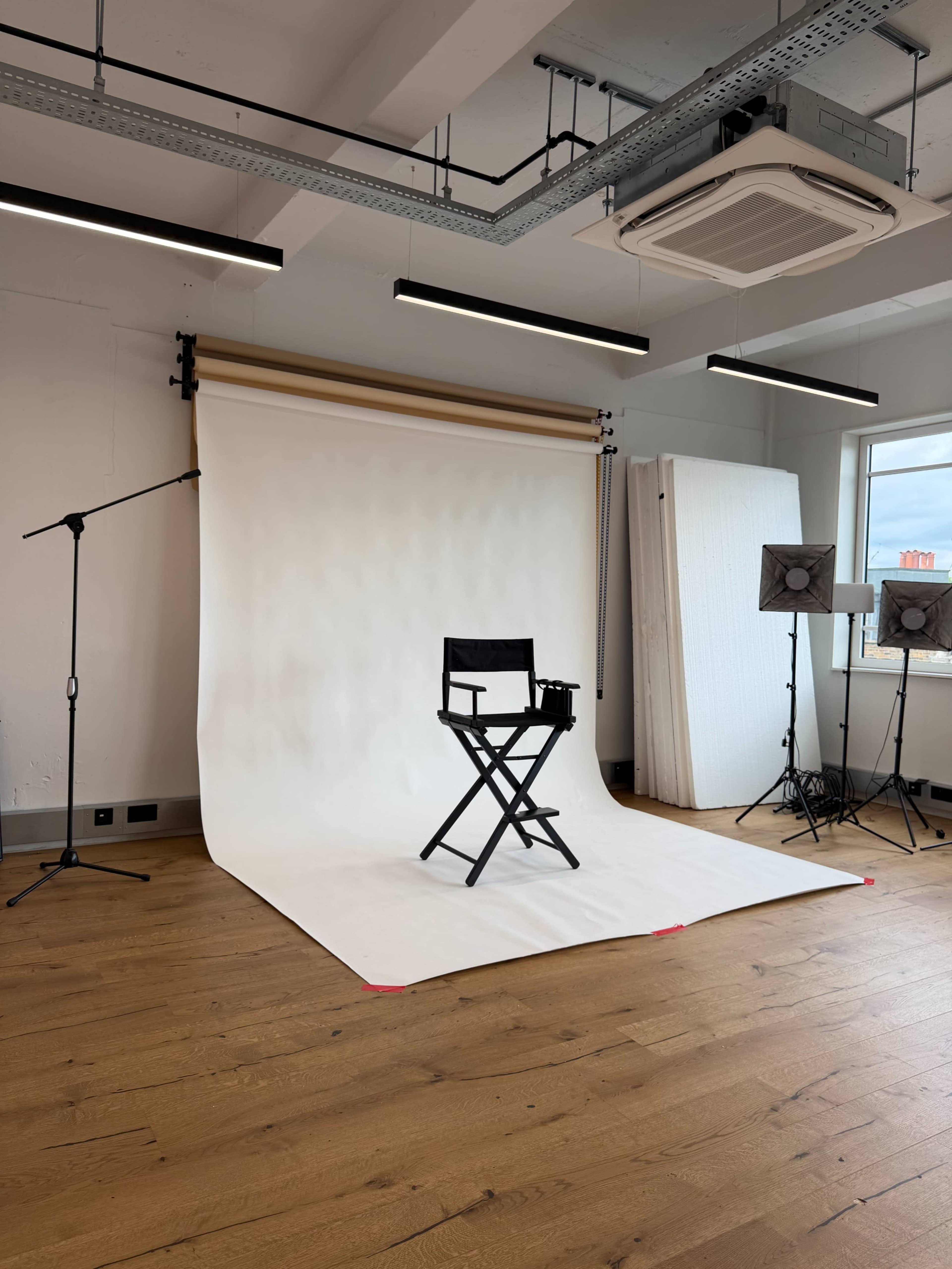 The image shows a photography studio with a white backdrop, a director's chair in the center, and lighting equipment positioned nearby.