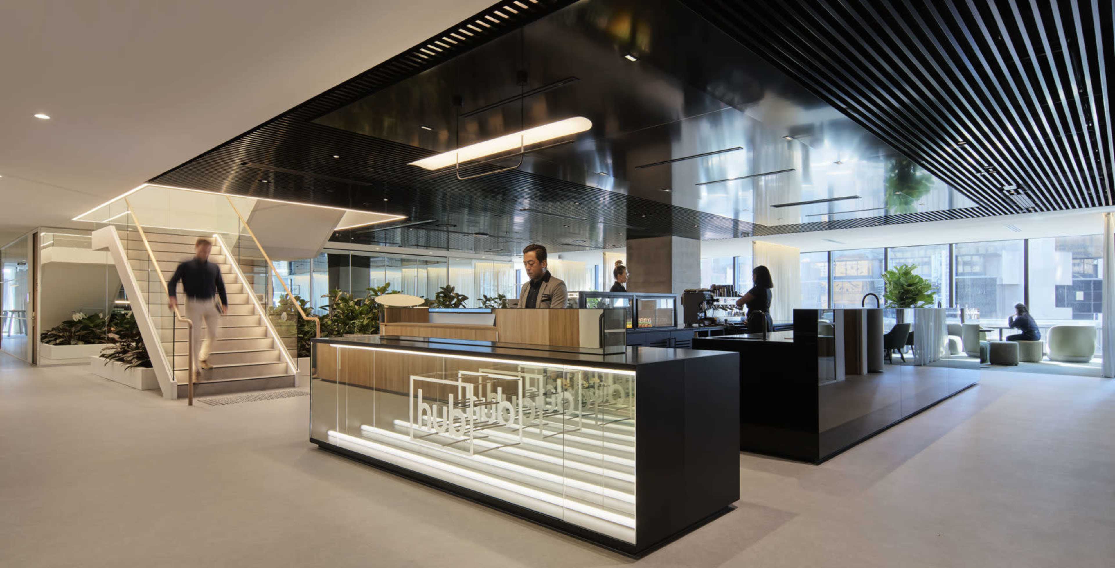 A modern office reception area with a sleek front desk, an open staircase, and abundant greenery throughout the space.