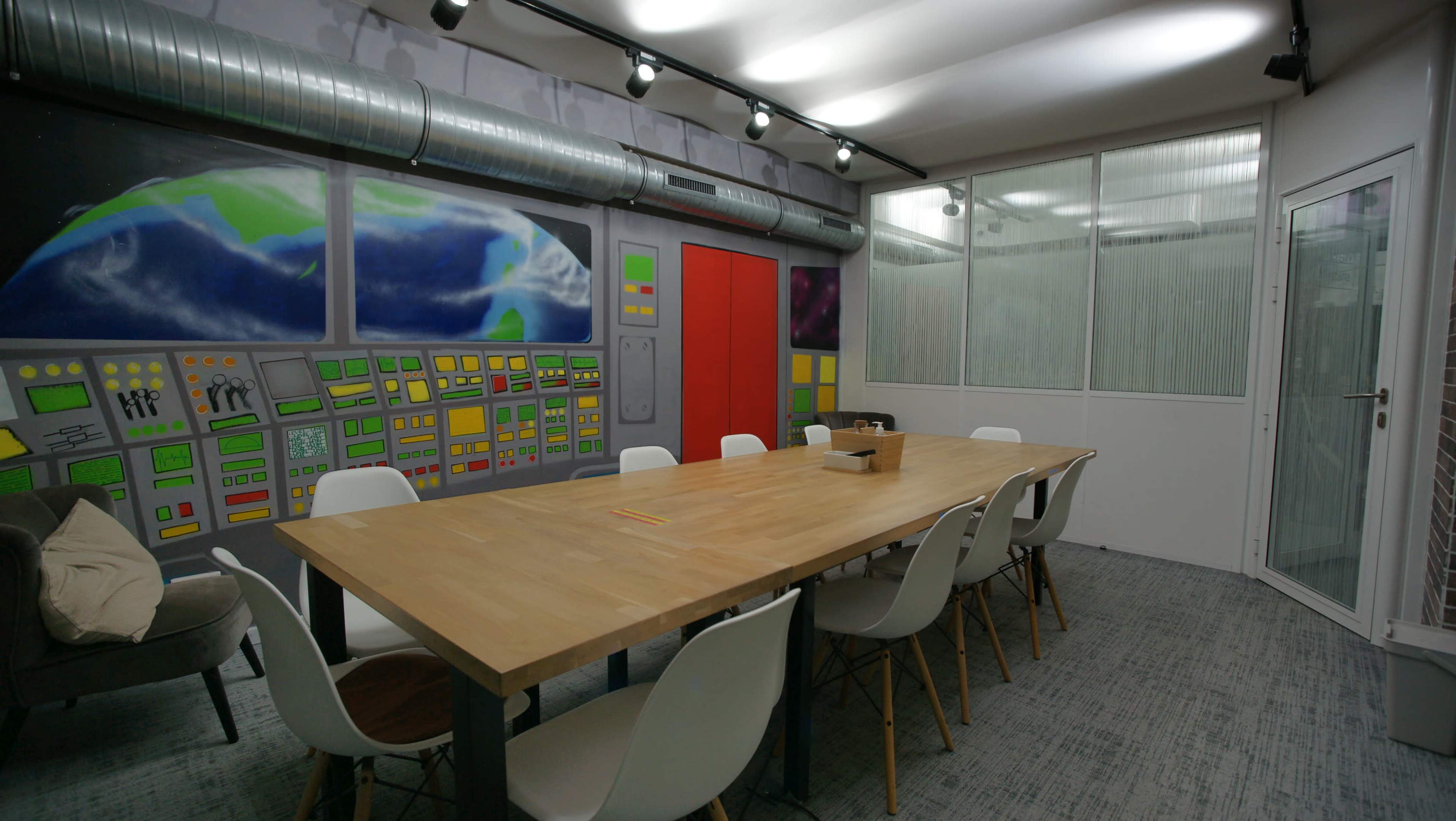 A modern conference room with a large wooden table surrounded by white chairs, featuring colorful murals on the walls and a glass-paneled area.