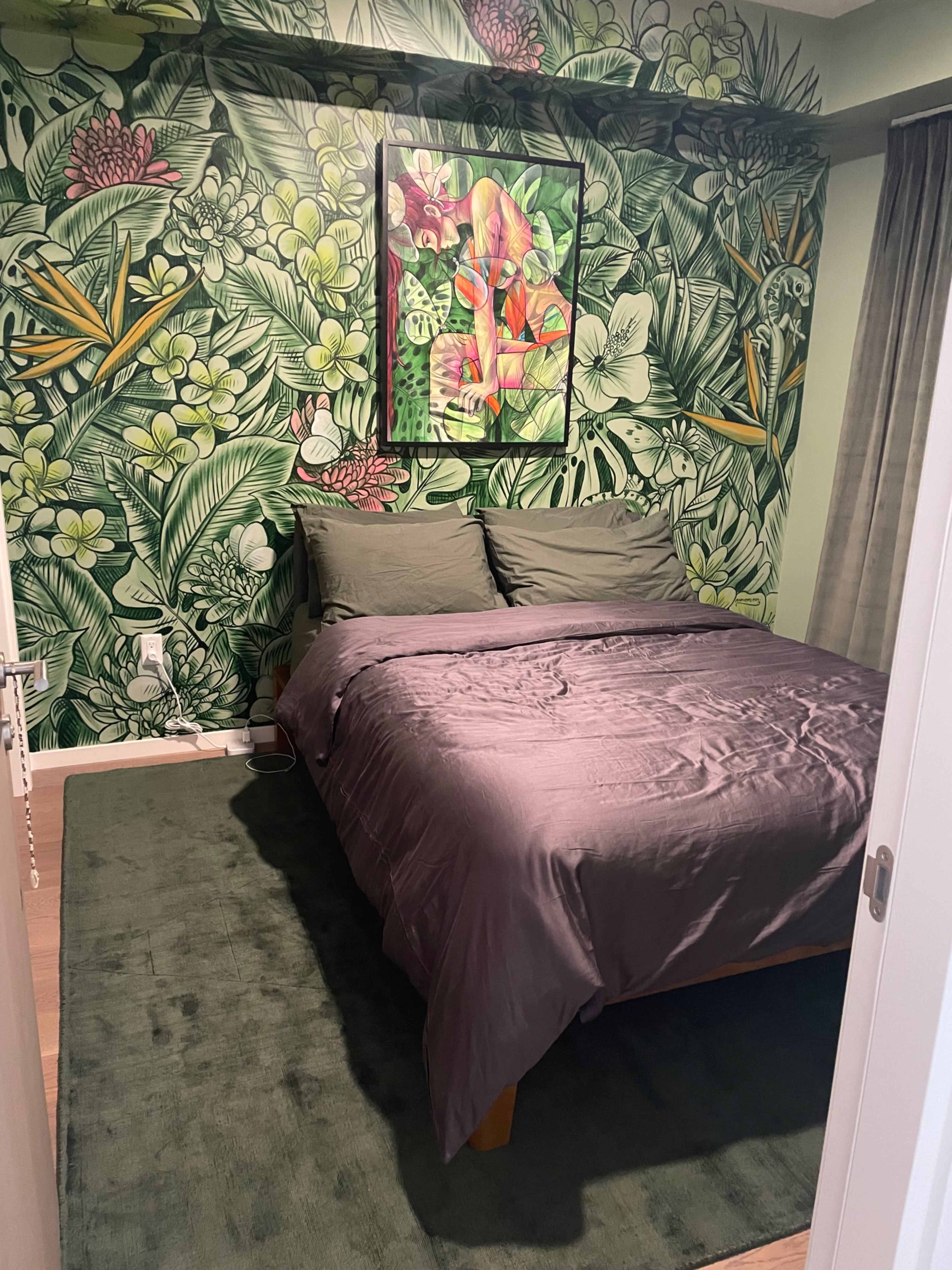 A small bedroom with a large bed covered in a gray comforter, surrounded by a green leafy wallpaper and a piece of artwork above the bed.
