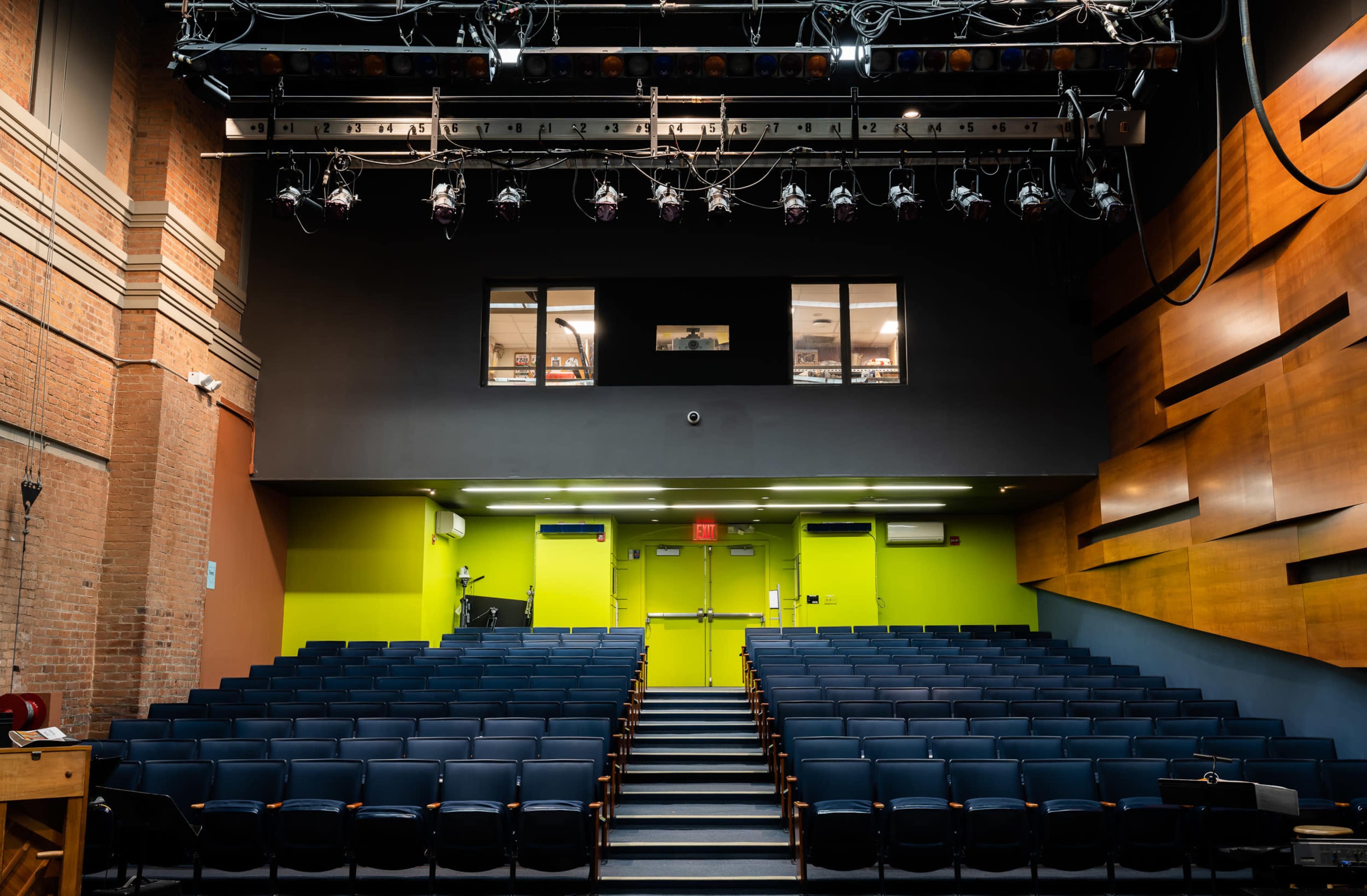 A theater auditorium with rows of blue seats facing a stage, overhead lighting, and green and brown walls.