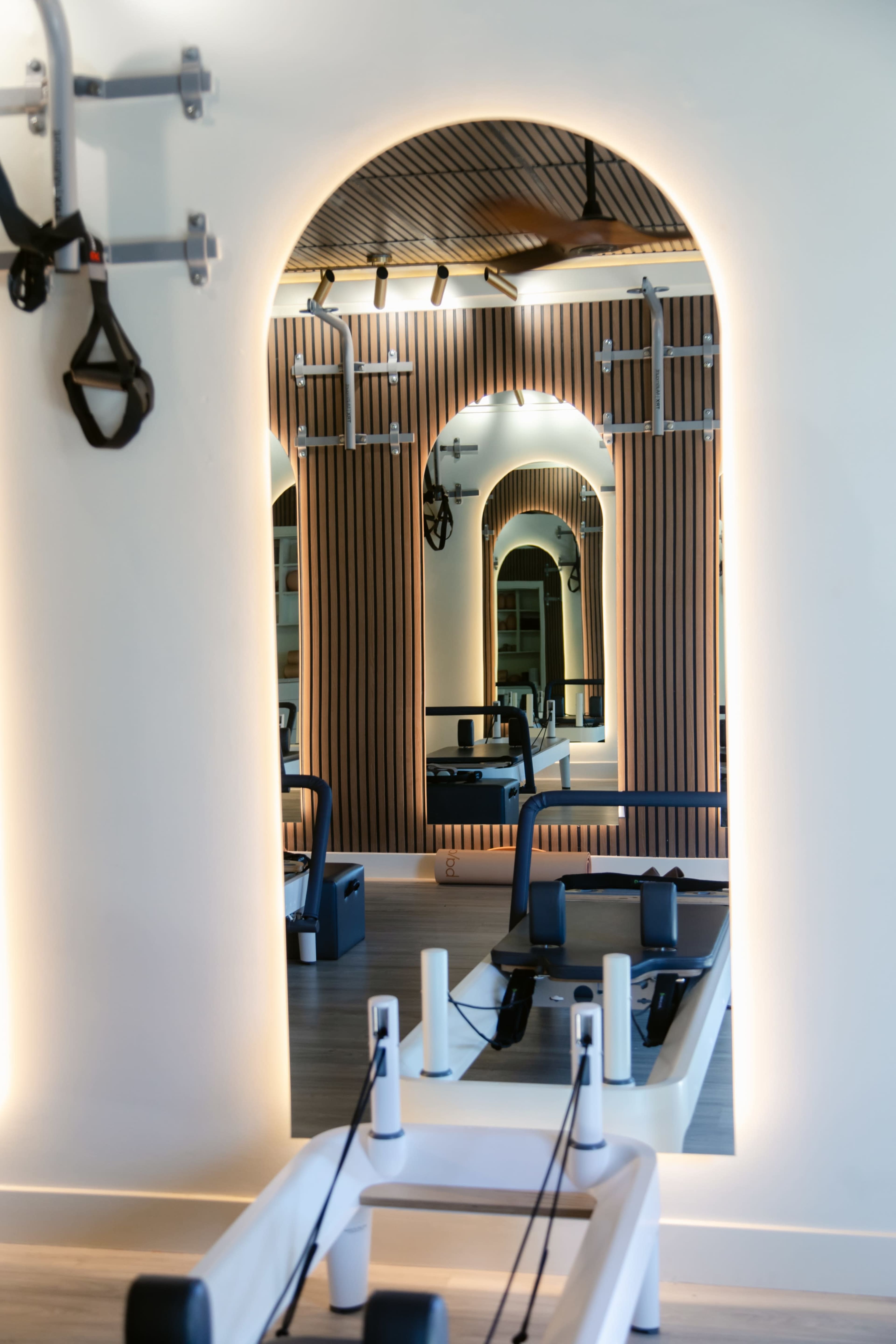 The image shows a modern fitness studio with mirrored walls, wooden paneling, and Pilates equipment arranged in the foreground.