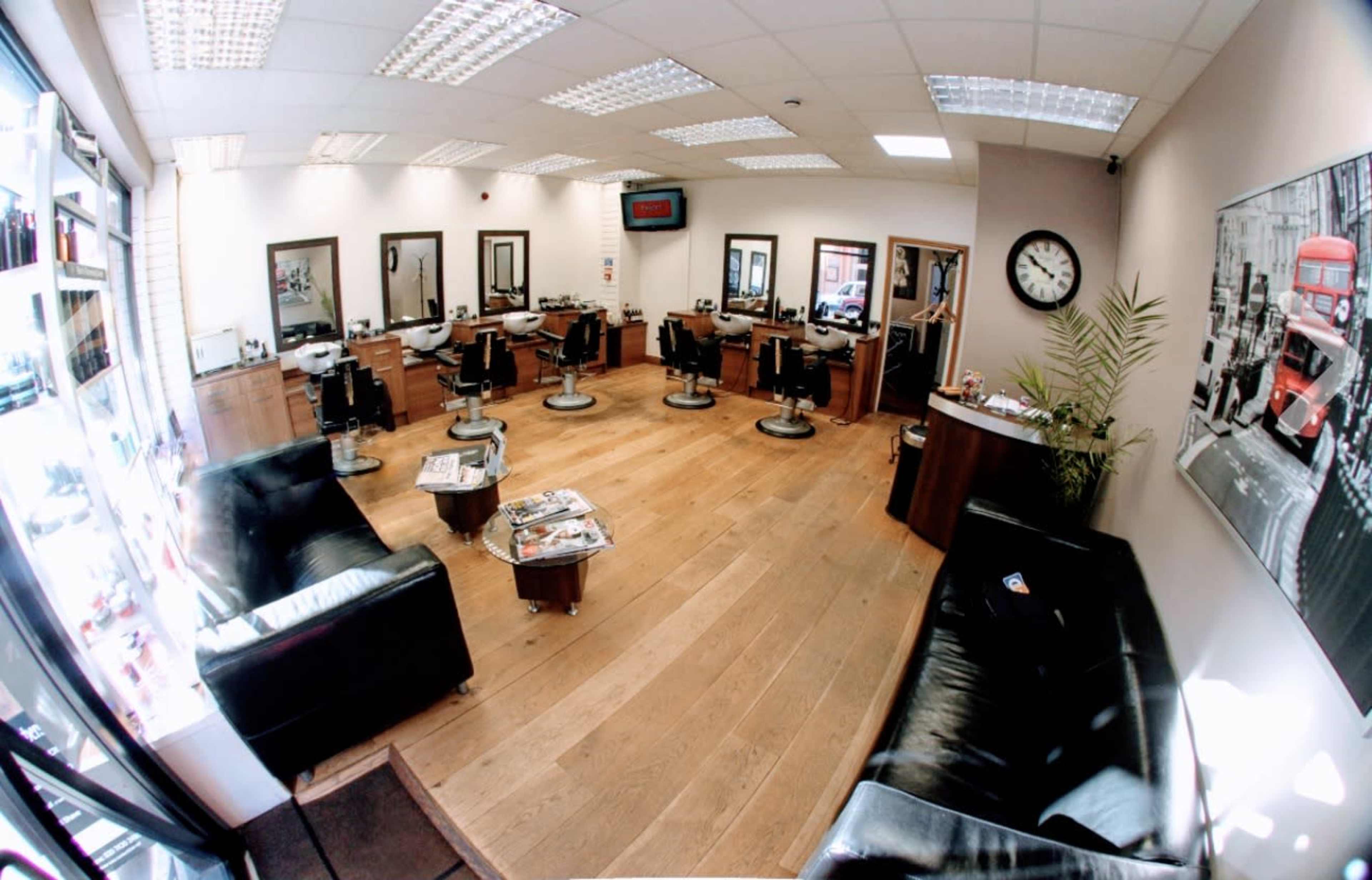 The image depicts a well-lit hair salon with several styling stations, black leather furniture, and a large wall clock.