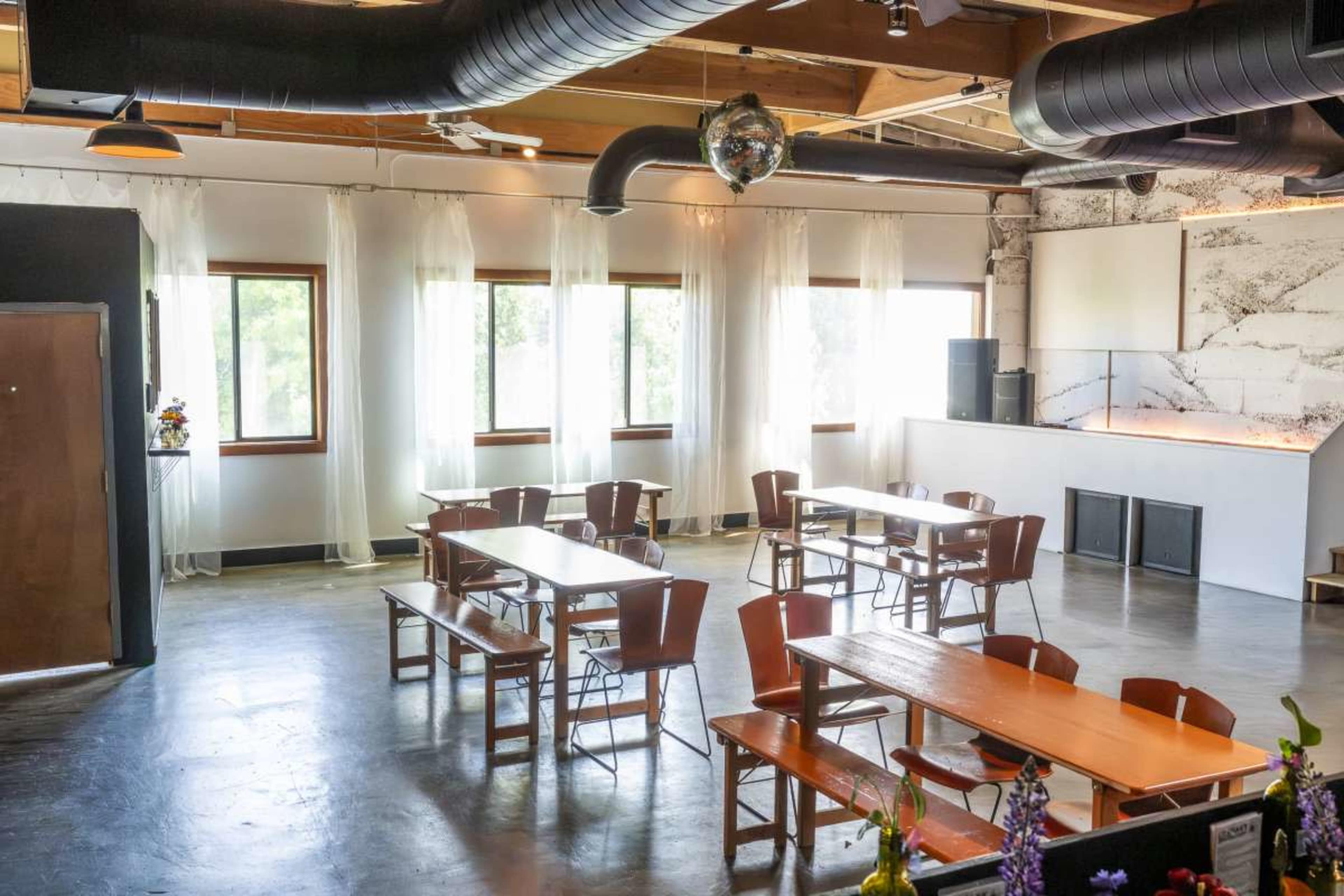 The image shows a spacious dining area with several wooden tables and benches arranged neatly, large windows allowing natural light to enter, and a minimalist counter in the background.