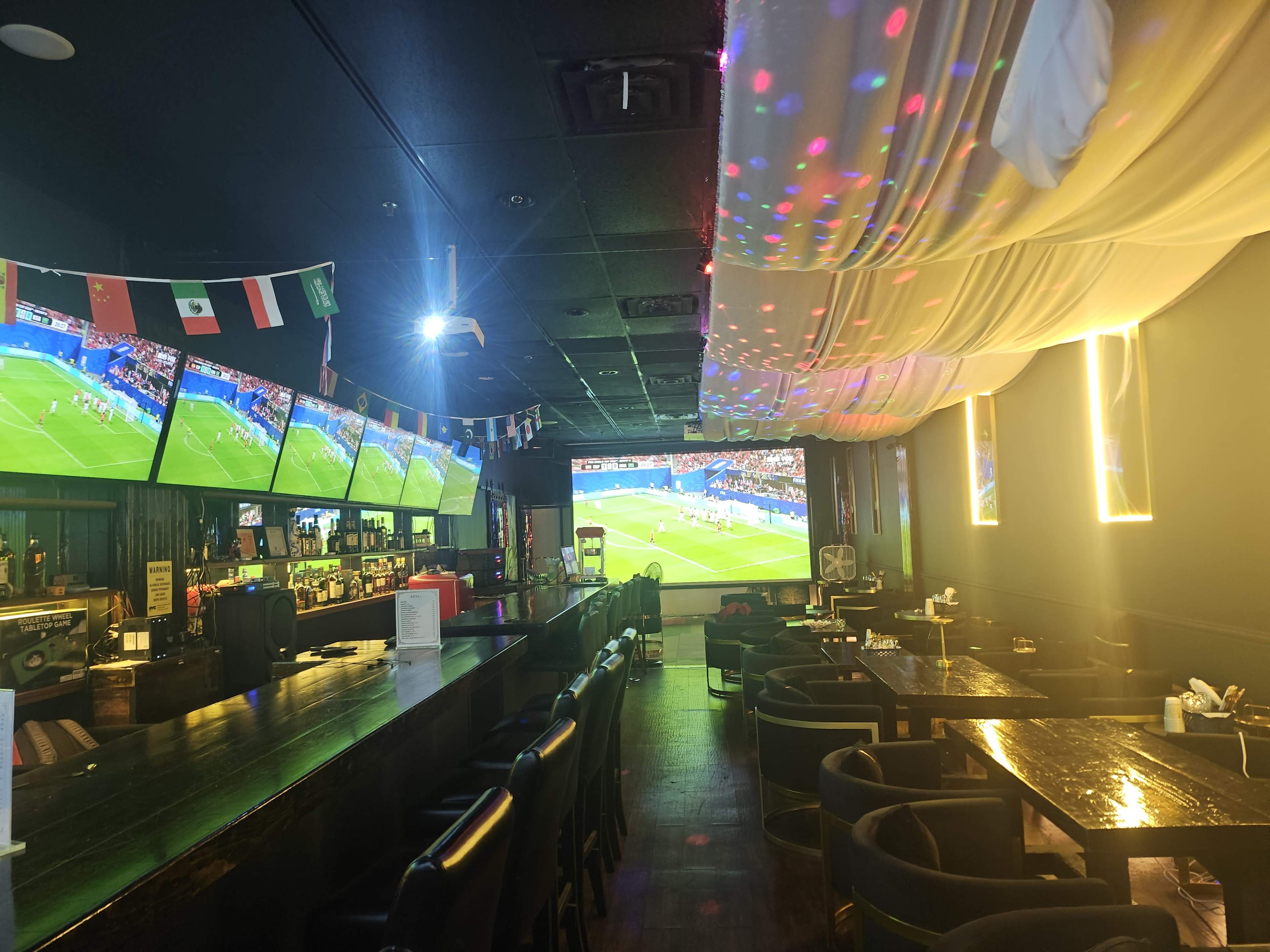 The image shows a dimly lit sports bar featuring multiple screens displaying a soccer match, with a long bar area and seating arranged in front of the screens.