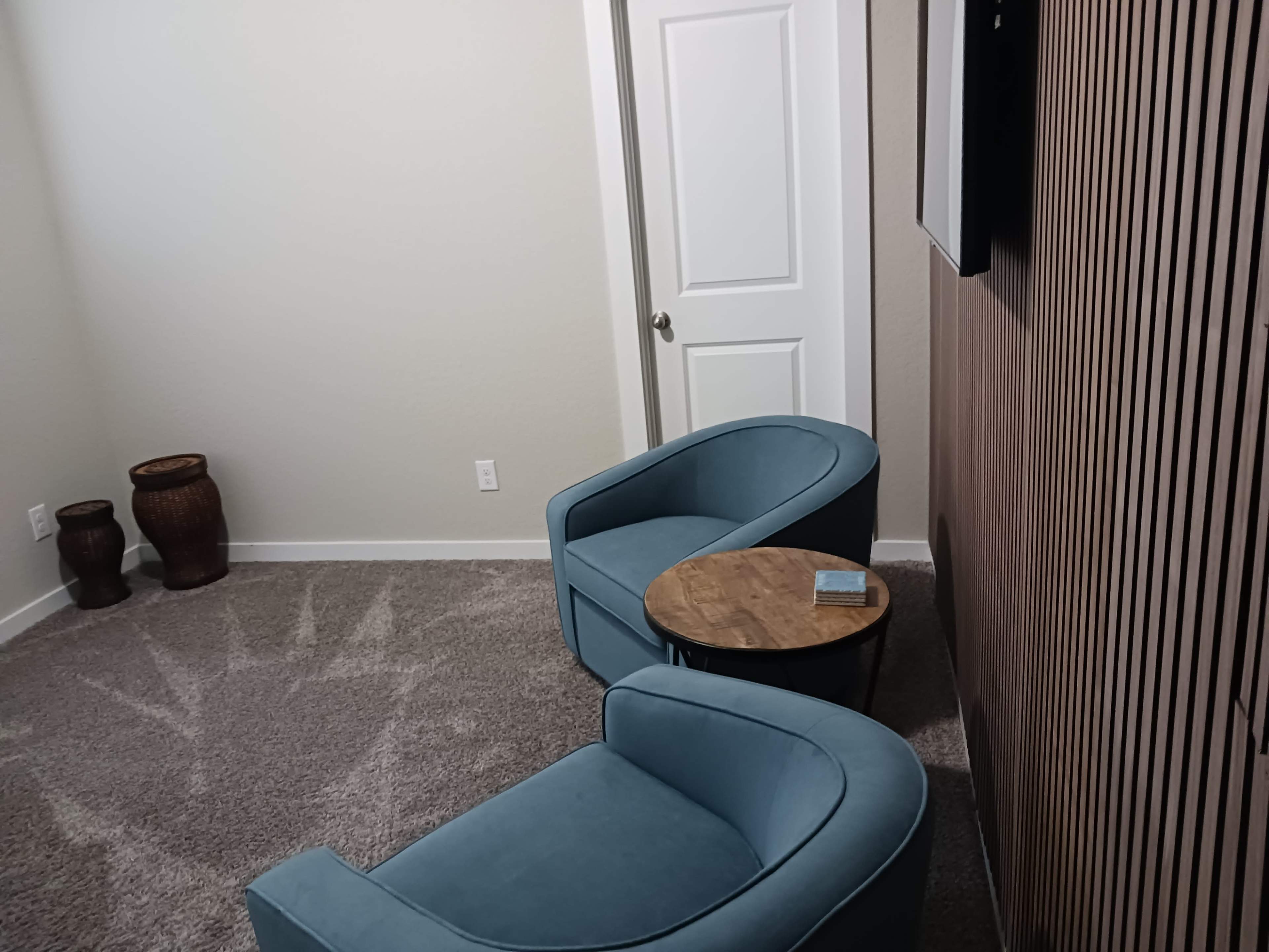 The image shows a small, cozy room with two blue chairs facing a wooden table, accompanied by decorative pots in the corner and a closed door in the background.