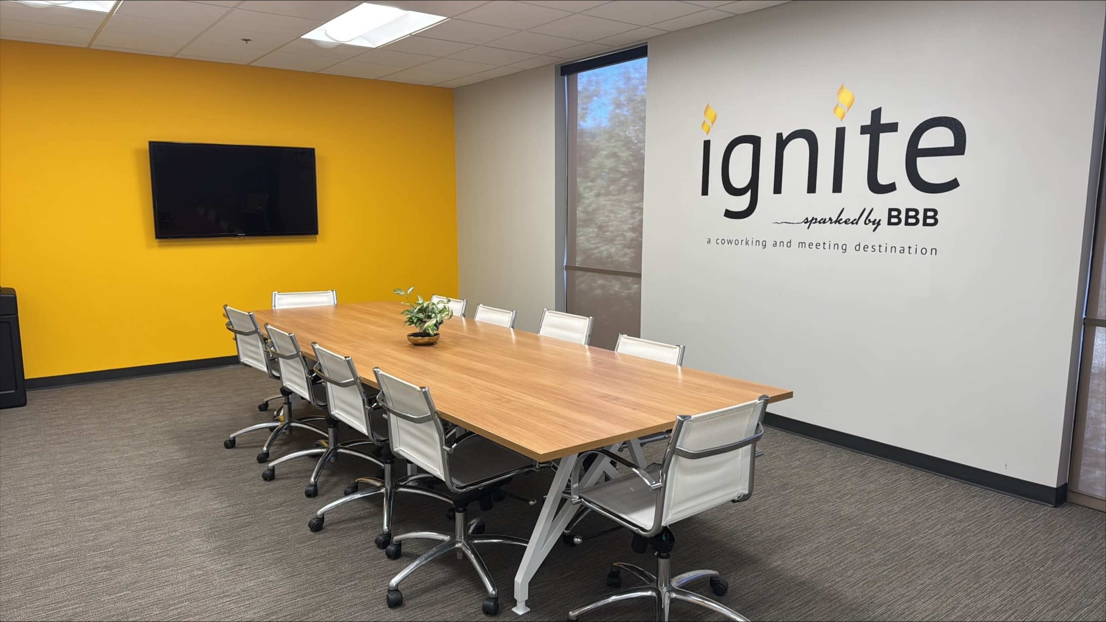 The image shows a modern conference room with a long wooden table surrounded by white chairs, a television mounted on the wall, and a bright yellow accent wall featuring the word "ignite."
