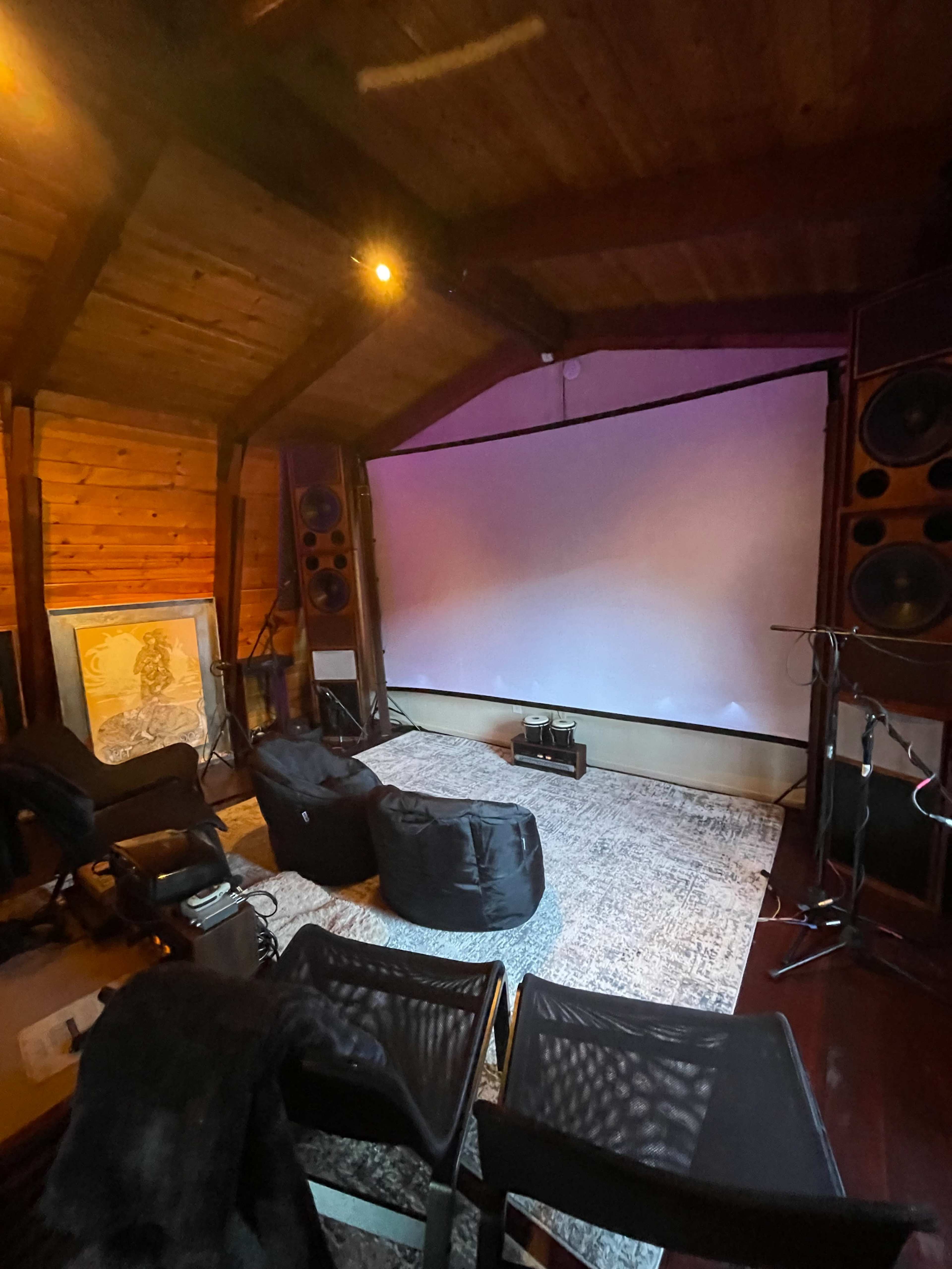 A cozy home theater setup featuring a large screen, speakers, bean bag chairs, and a rug in a wooden room.