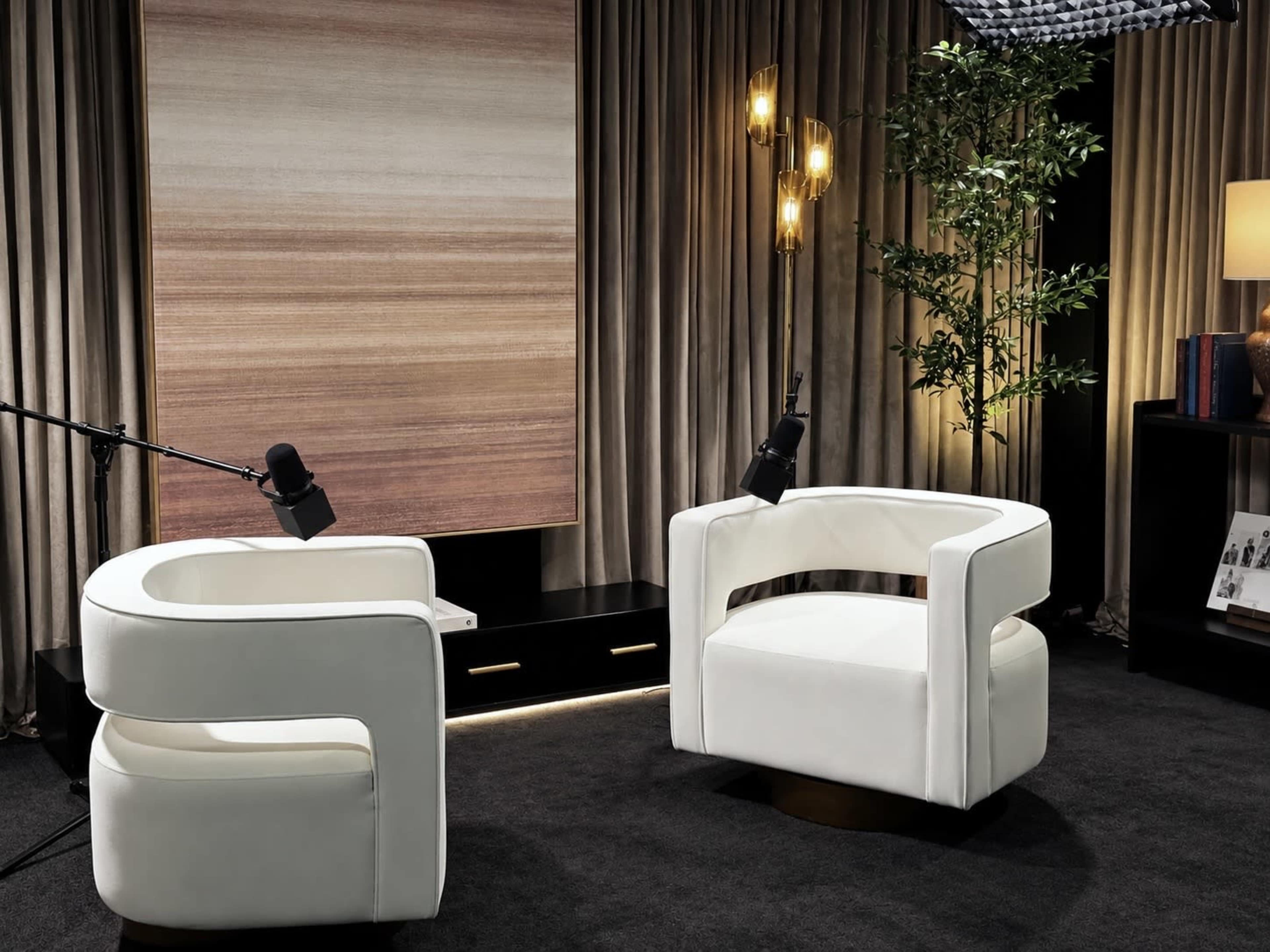 The image features two modern white chairs arranged facing each other in a well-lit interior space, with a textured backdrop and decorative elements like plants and lighting fixtures.