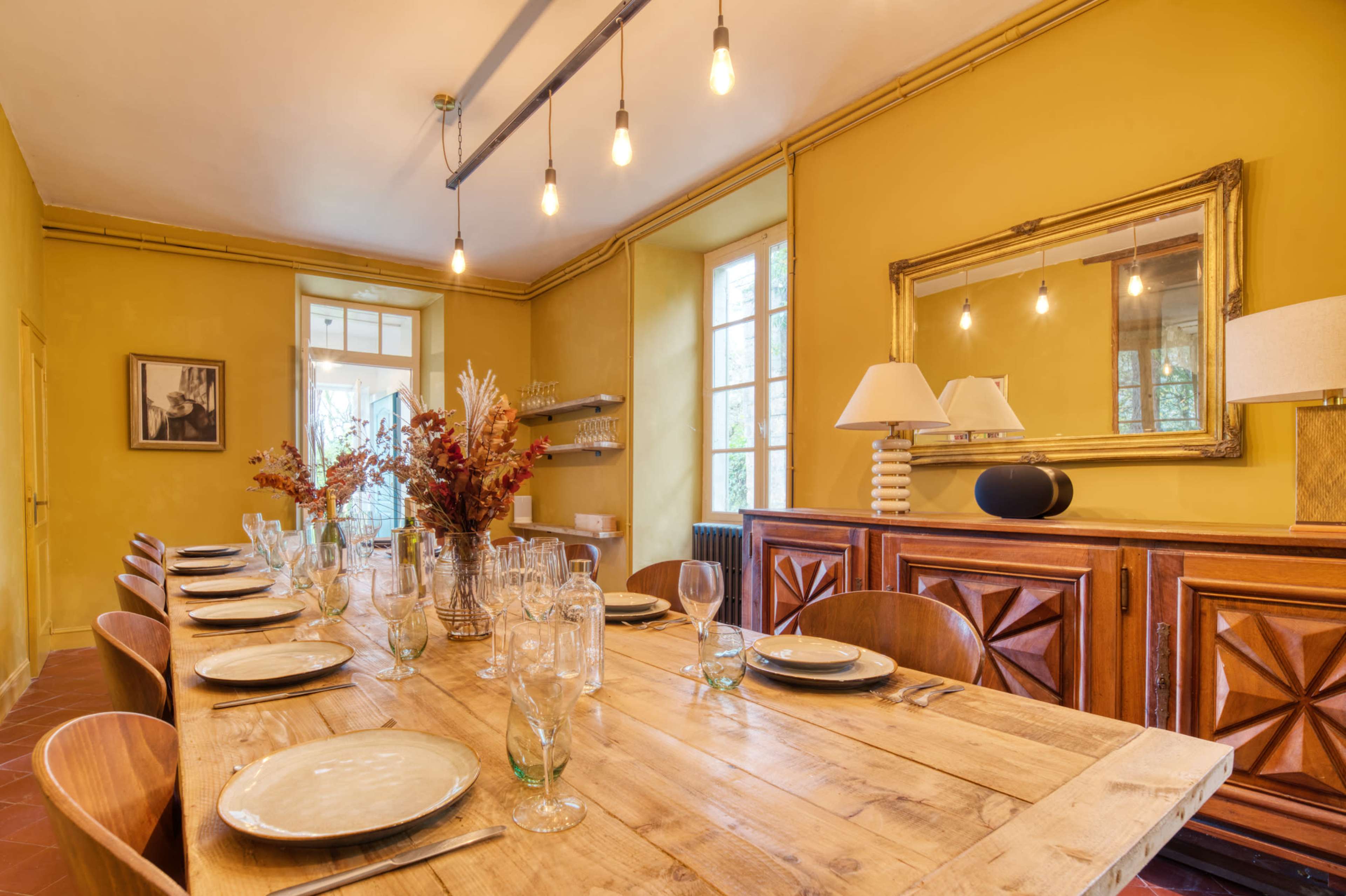 A long wooden dining table is set with plates and glassware in a sunny room featuring yellow walls and pendant lighting.