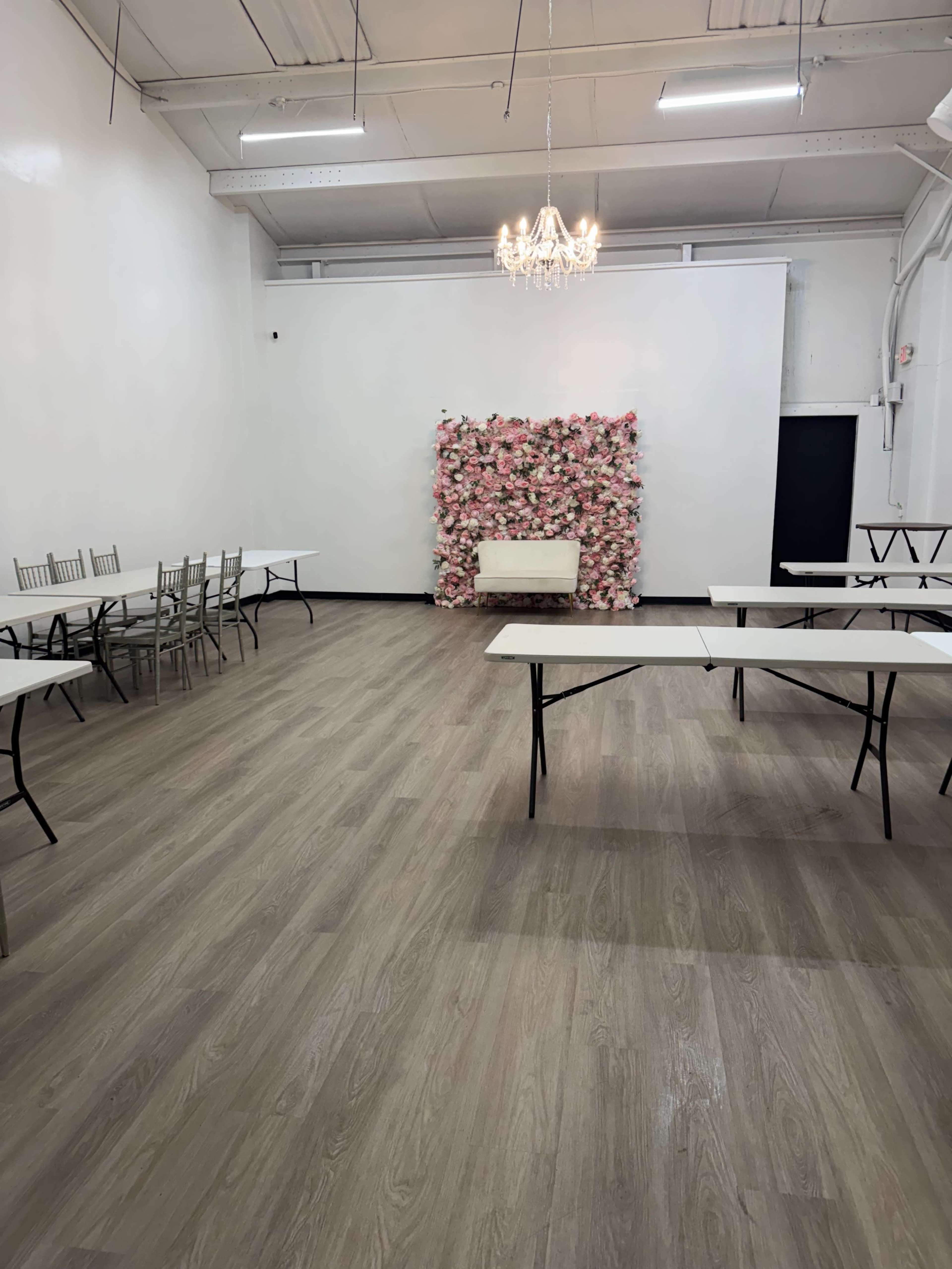 The image shows a spacious event room with several tables arranged around, featuring a floral backdrop and a chandelier overhead.