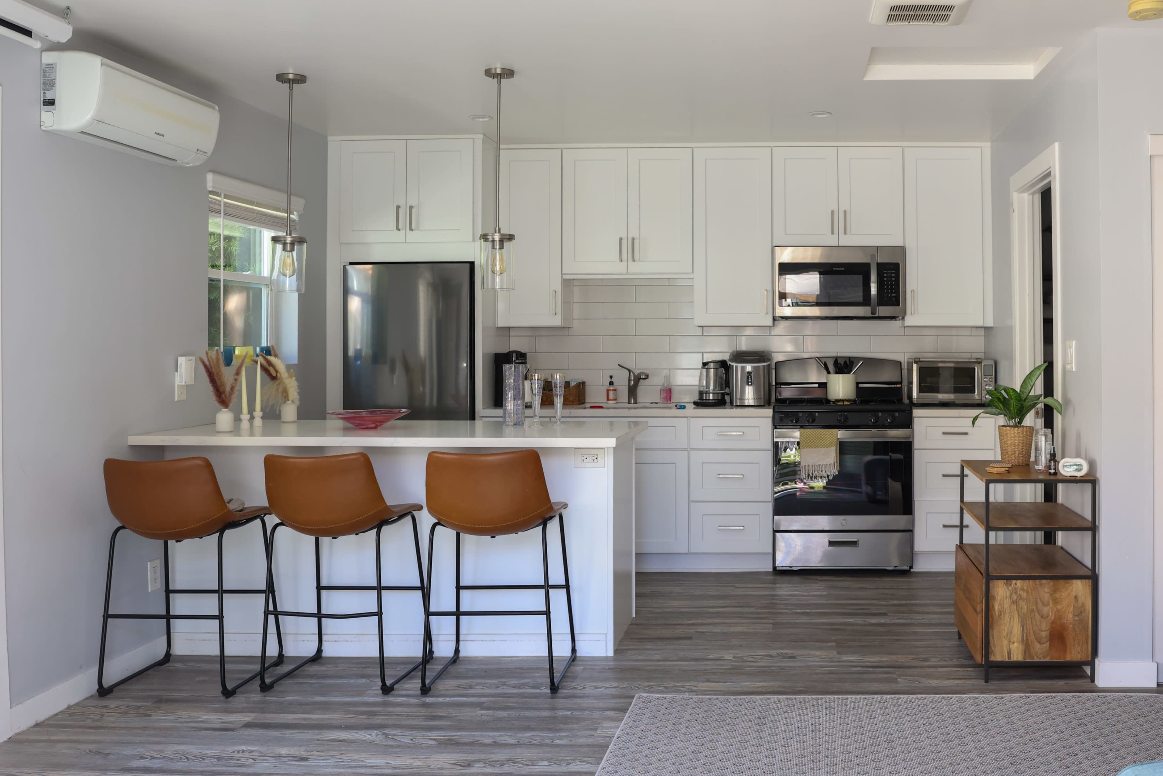 The image shows a modern kitchen with white cabinets, stainless steel appliances, and a countertop with three brown bar stools.