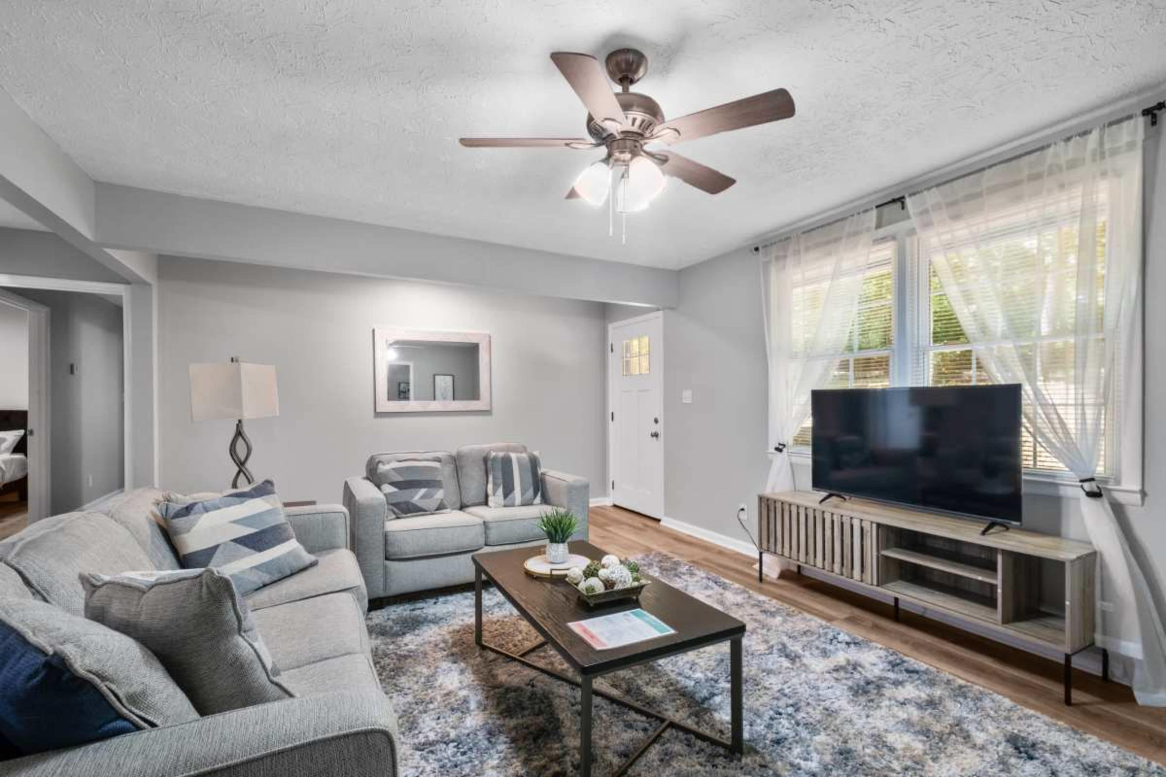 The living room features a light gray color scheme, a ceiling fan, a television on a wooden stand, and a coffee table with decorative items.