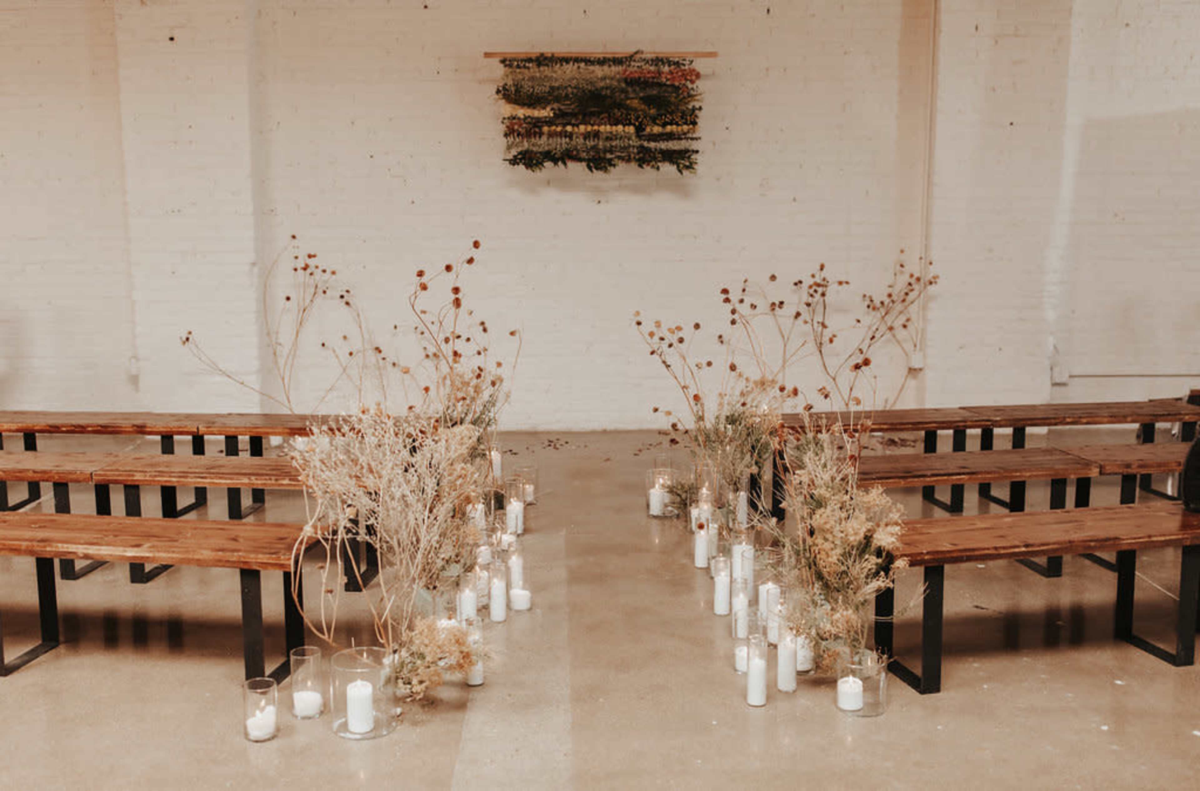 A minimalist ceremony setup with wooden benches arranged in two rows, flanking a central aisle lined with dried flowers and candles.