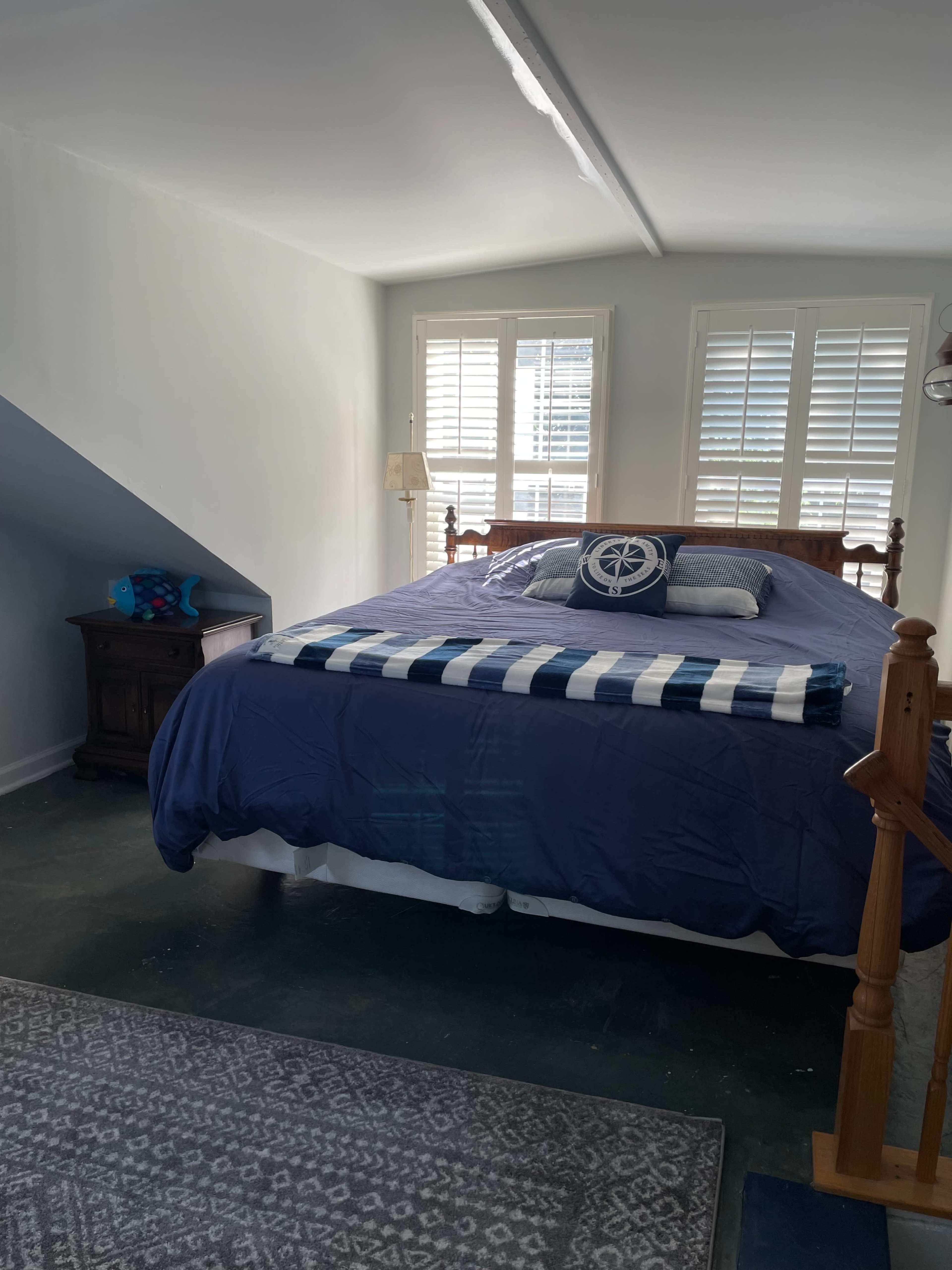 A bedroom features a large bed with a navy blue comforter, two pillows, and a decorative throw, beside a wooden nightstand and under a staircase, with window shutters letting in natural light.