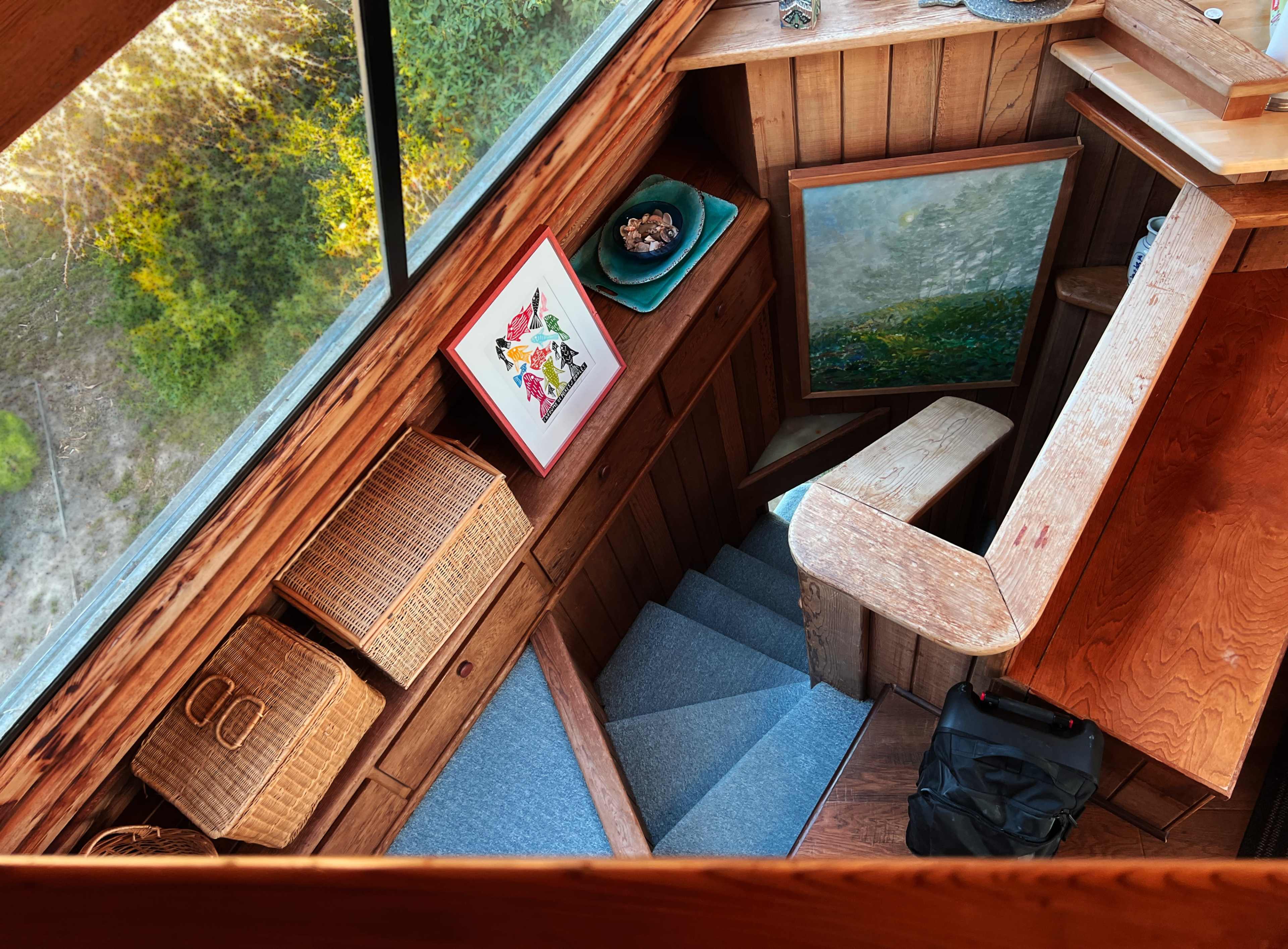 The image shows a wooden stairwell with a small landing, decorated with woven baskets, a framed art piece, and a view of greenery through a large window.