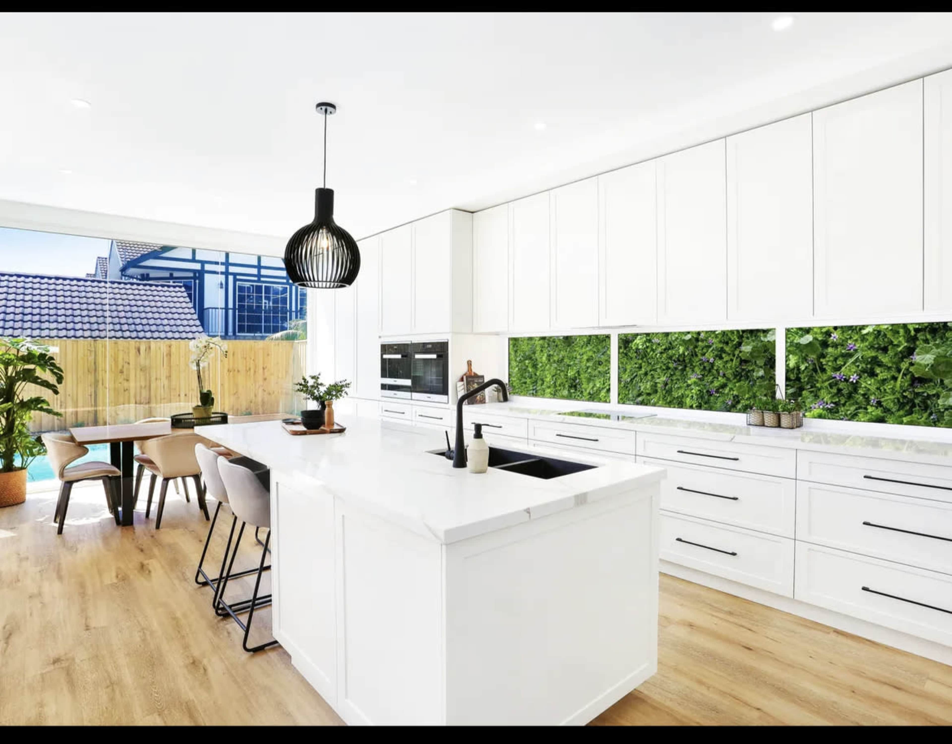 A modern kitchen features a central island with a sink, surrounded by sleek cabinetry and natural greenery visible through large windows.