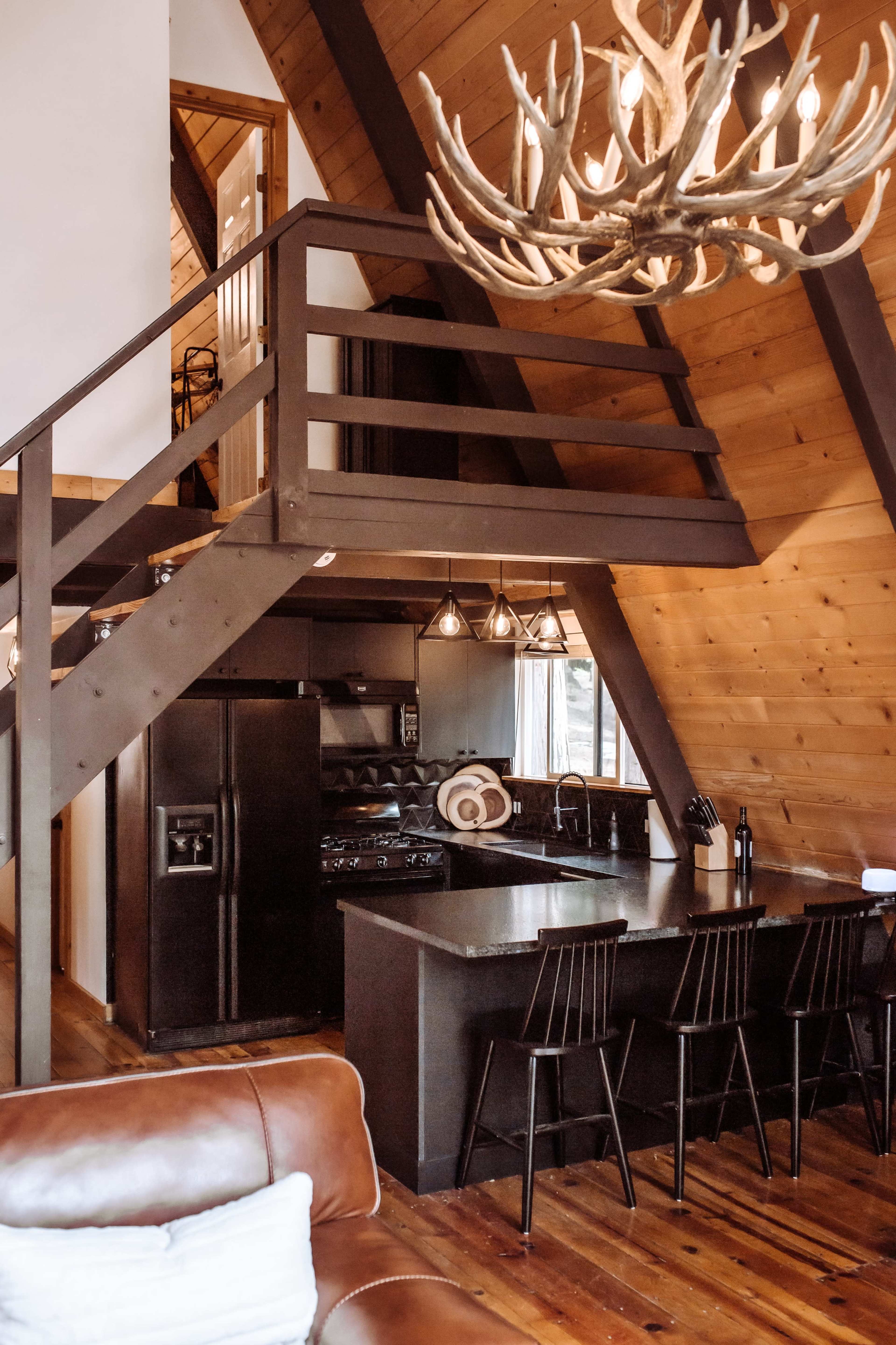 The image shows a modern cabin interior featuring an open kitchen with a black countertop, a dining area with black bar stools, and a loft area illuminated by a large antler chandelier.