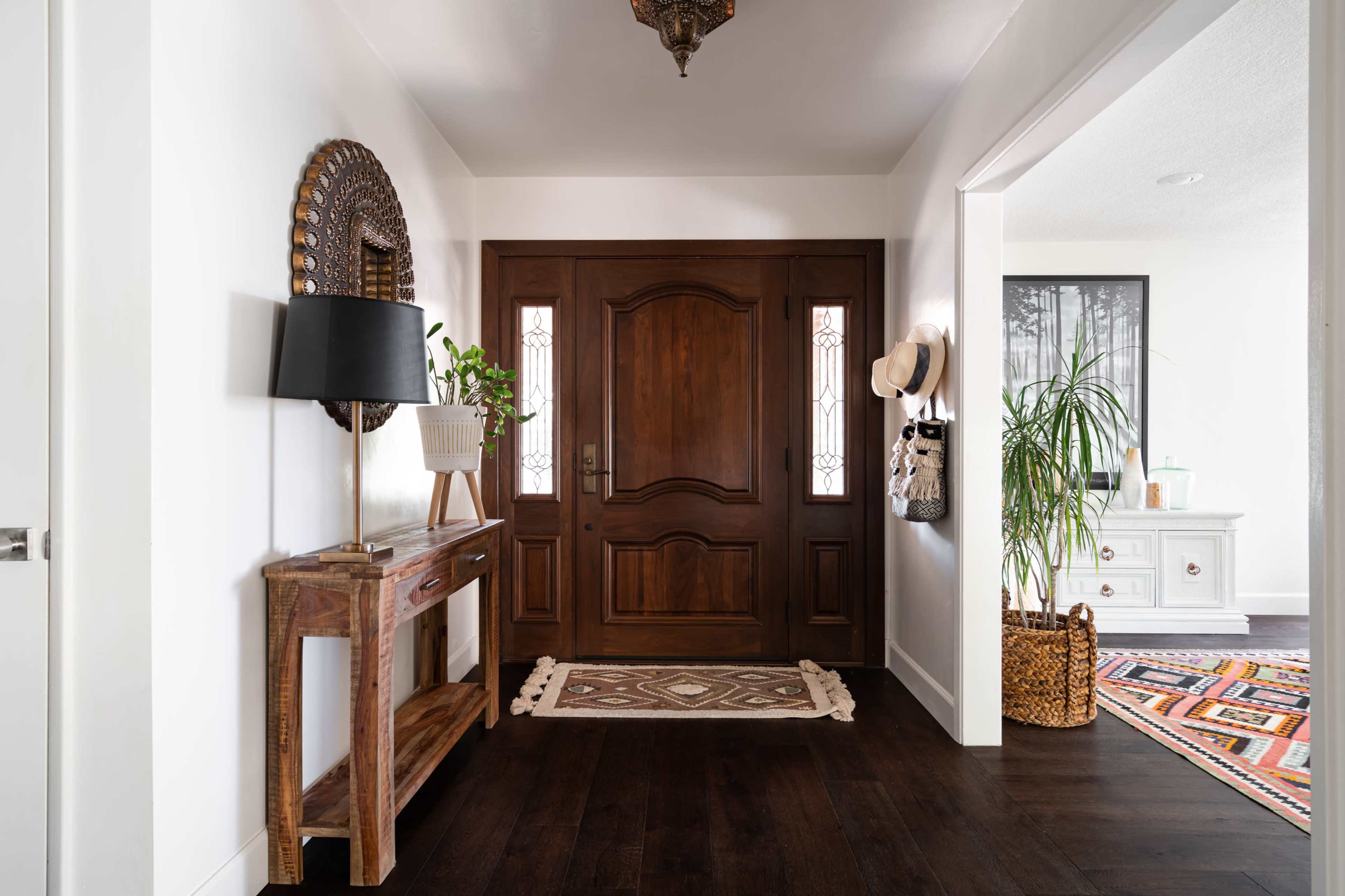 A modern entryway with a wooden front door, a console table with a lamp and plant, and a patterned rug on dark hardwood flooring.
