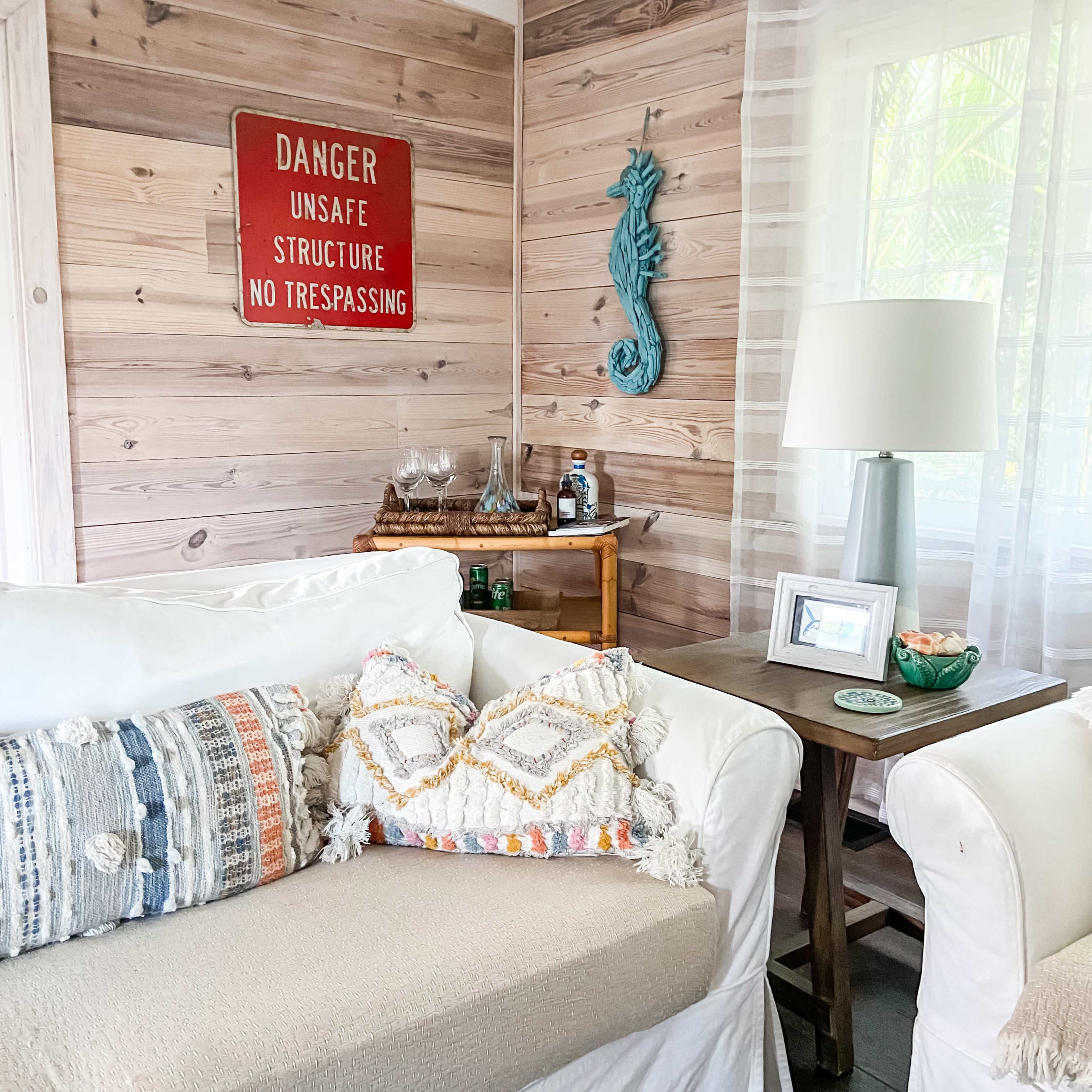The image shows a cozy living space featuring a white couch adorned with decorative pillows, a wooden side table with a lamp, and a wall with a vintage "Danger Unsafe Structure No Trespassing" sign.