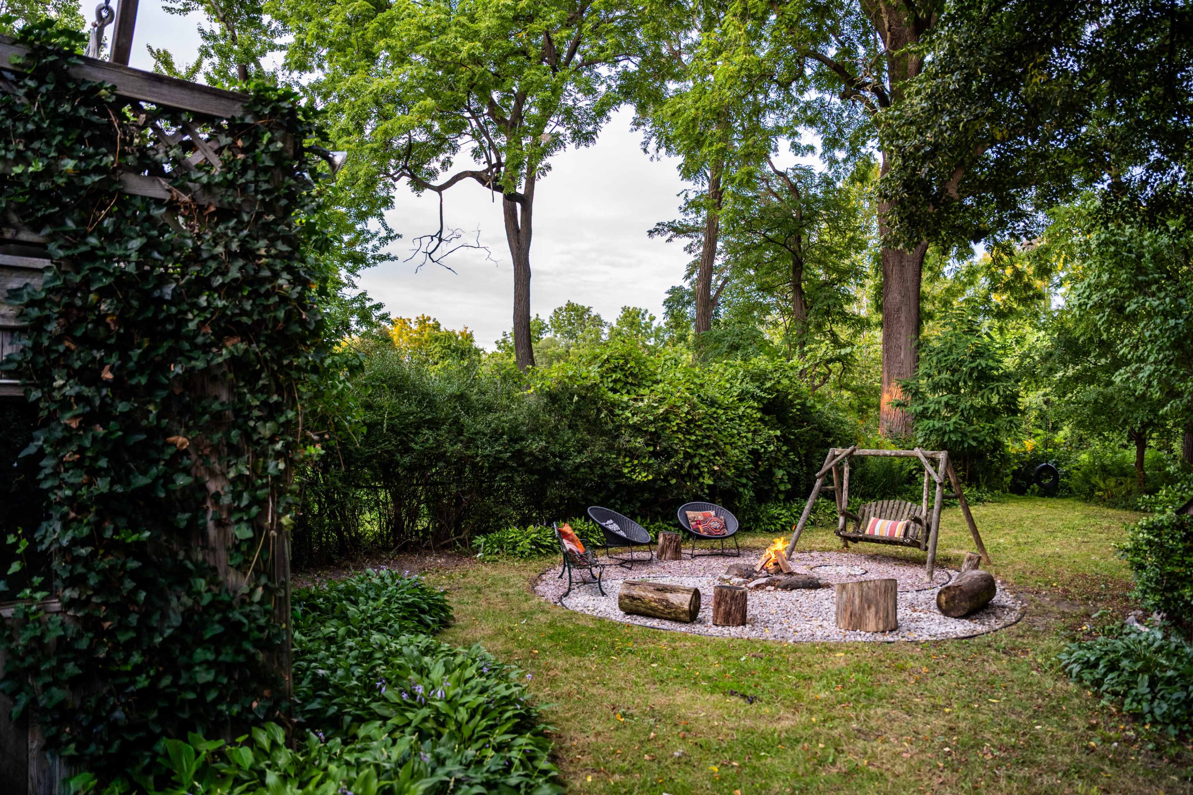 The image shows a backyard with a fire pit surrounded by logs and two chairs, set among trees and greenery.