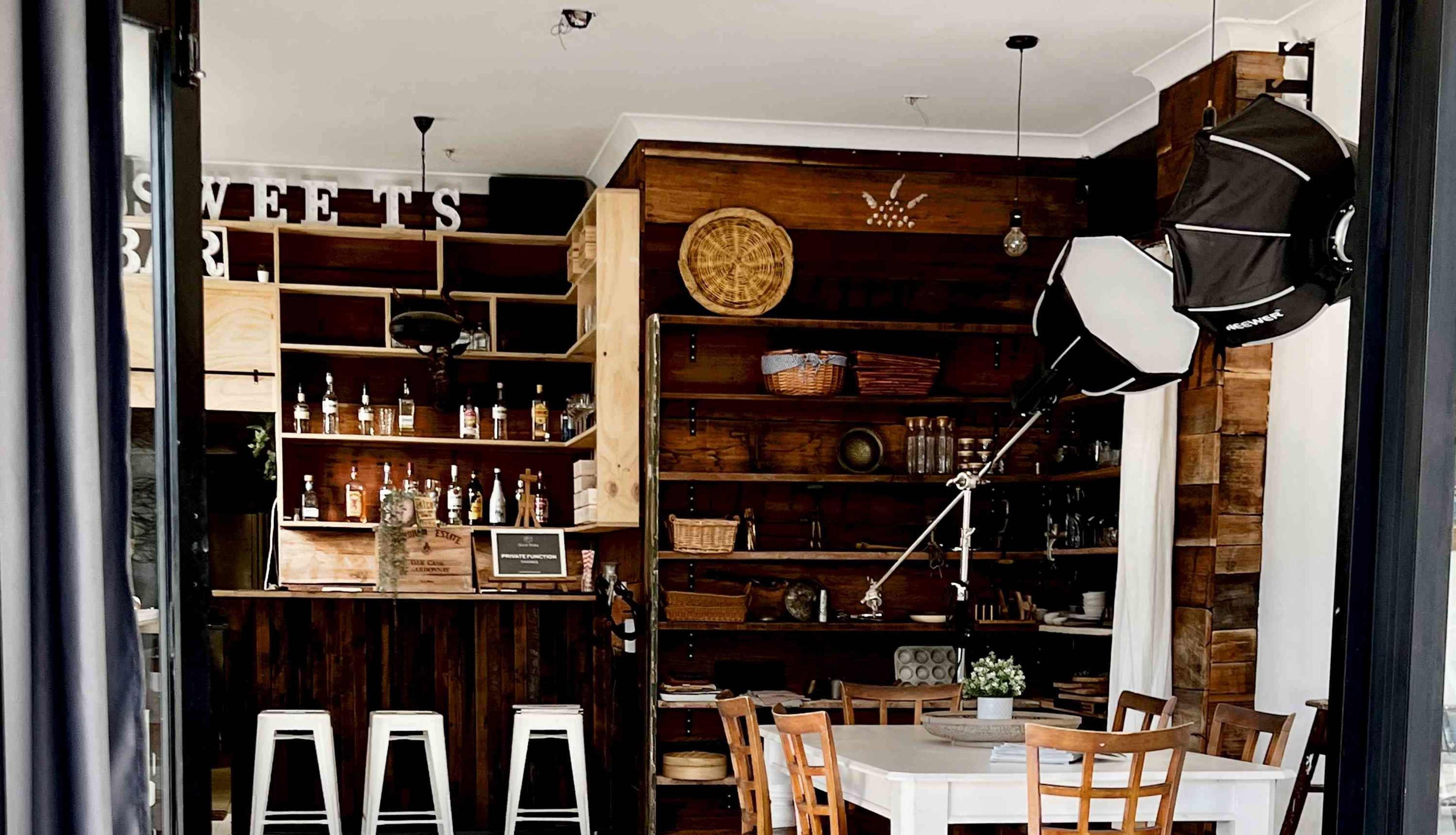 The image shows a rustic bar area with wooden shelves displaying bottles and decorative items, accompanied by a white dining table and chairs.
