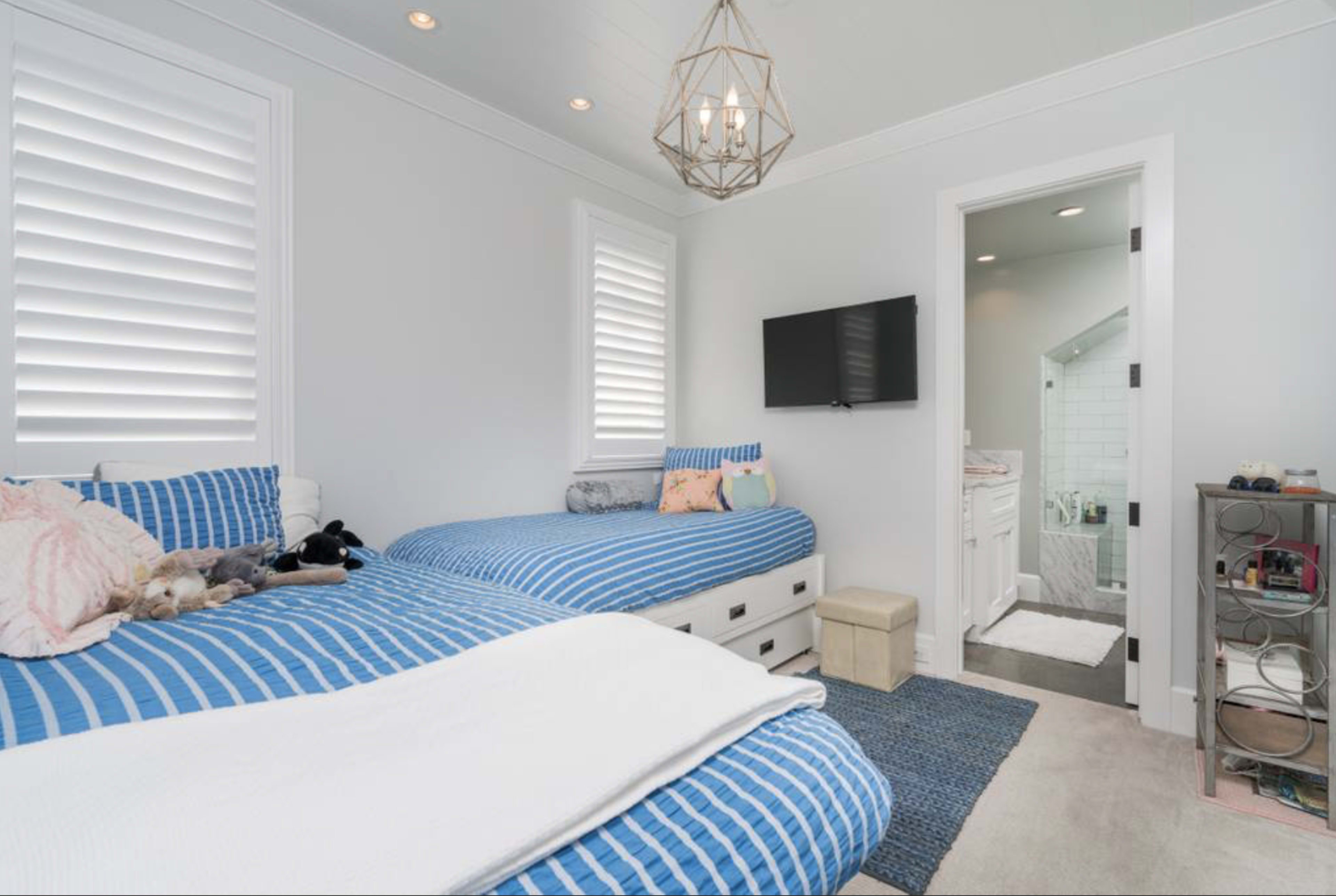 The image shows a well-lit bedroom with two beds featuring blue striped bedding, a television mounted on the wall, and an attached bathroom visible through an open door.