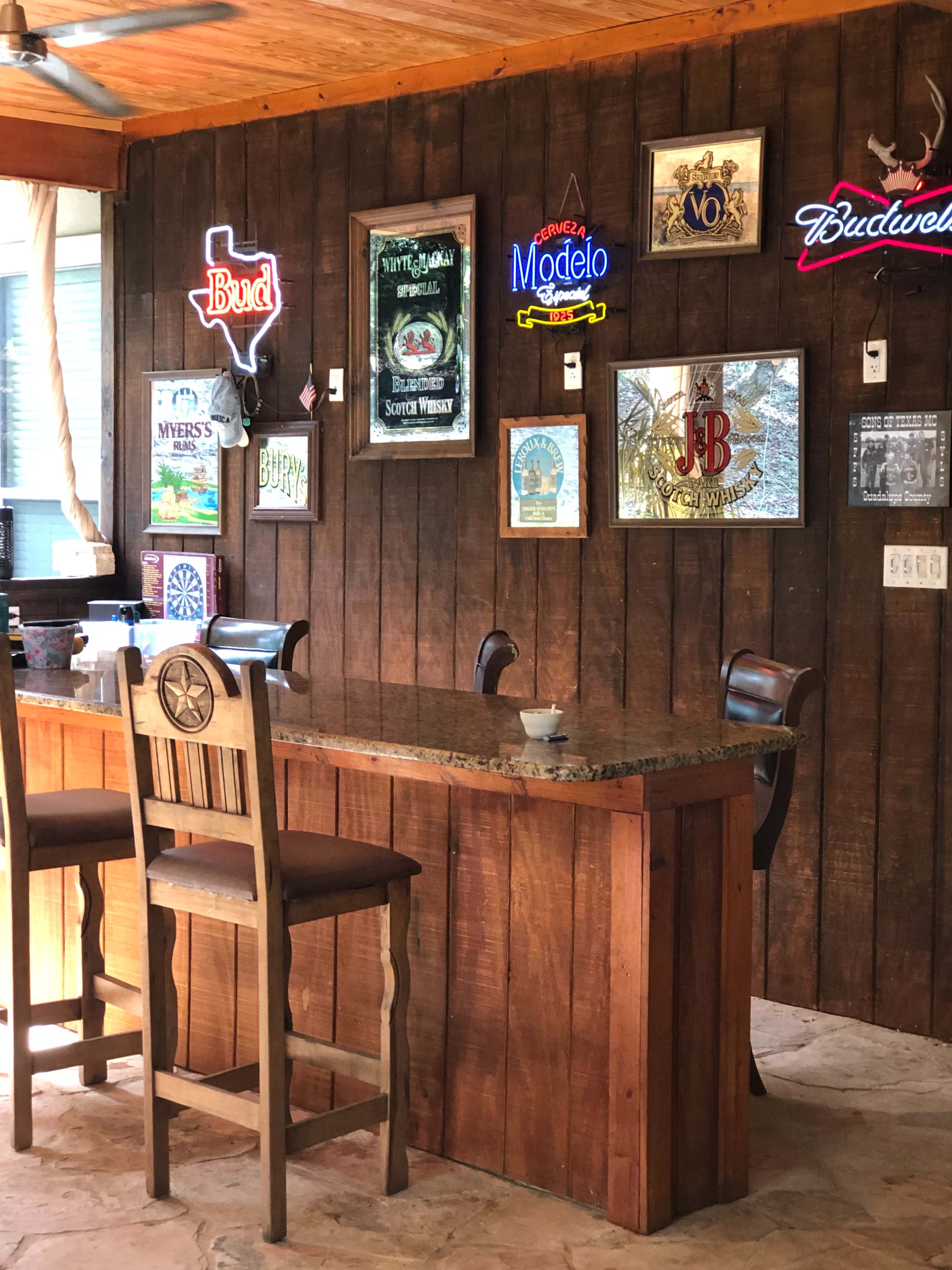 The image shows a wooden bar with high stools and a wall adorned with various neon signs and framed artwork.