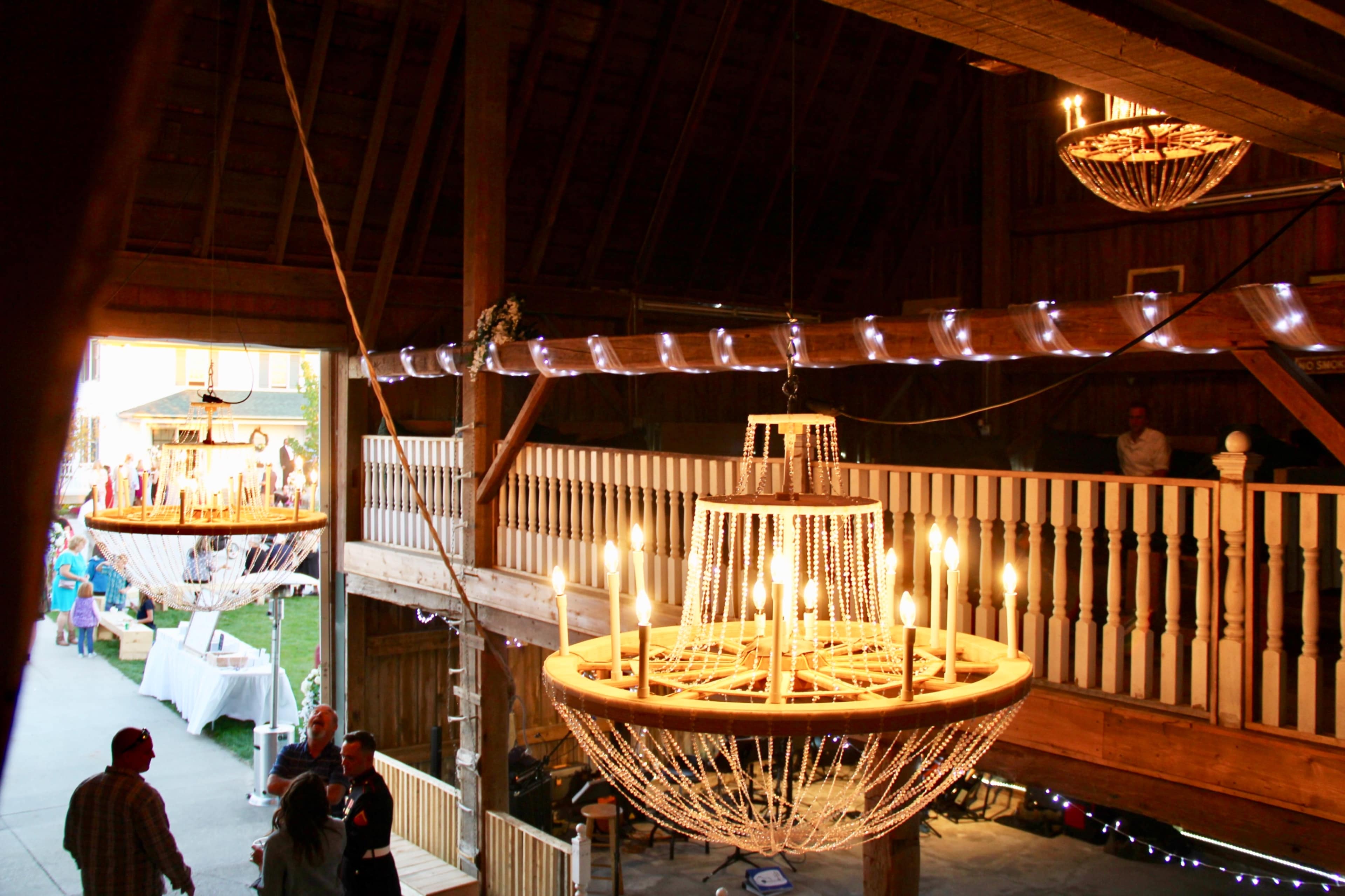 The interior of a barn features two large chandeliers and a balcony, with people gathered below and decorative lights overhead.
